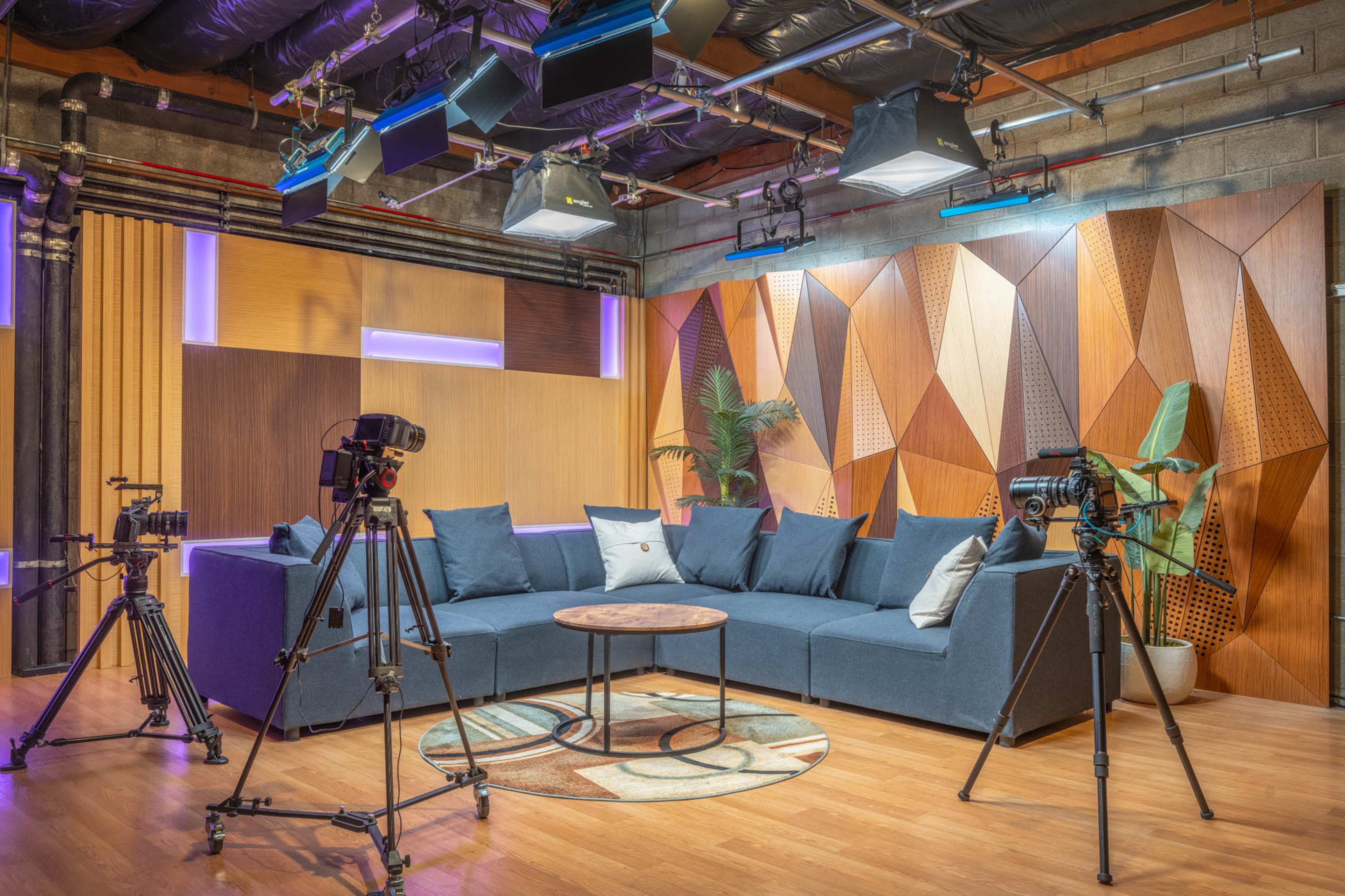 The image shows a well-lit studio setup featuring a plush sofa surrounded by multiple cameras and a round wooden table on a circular rug.