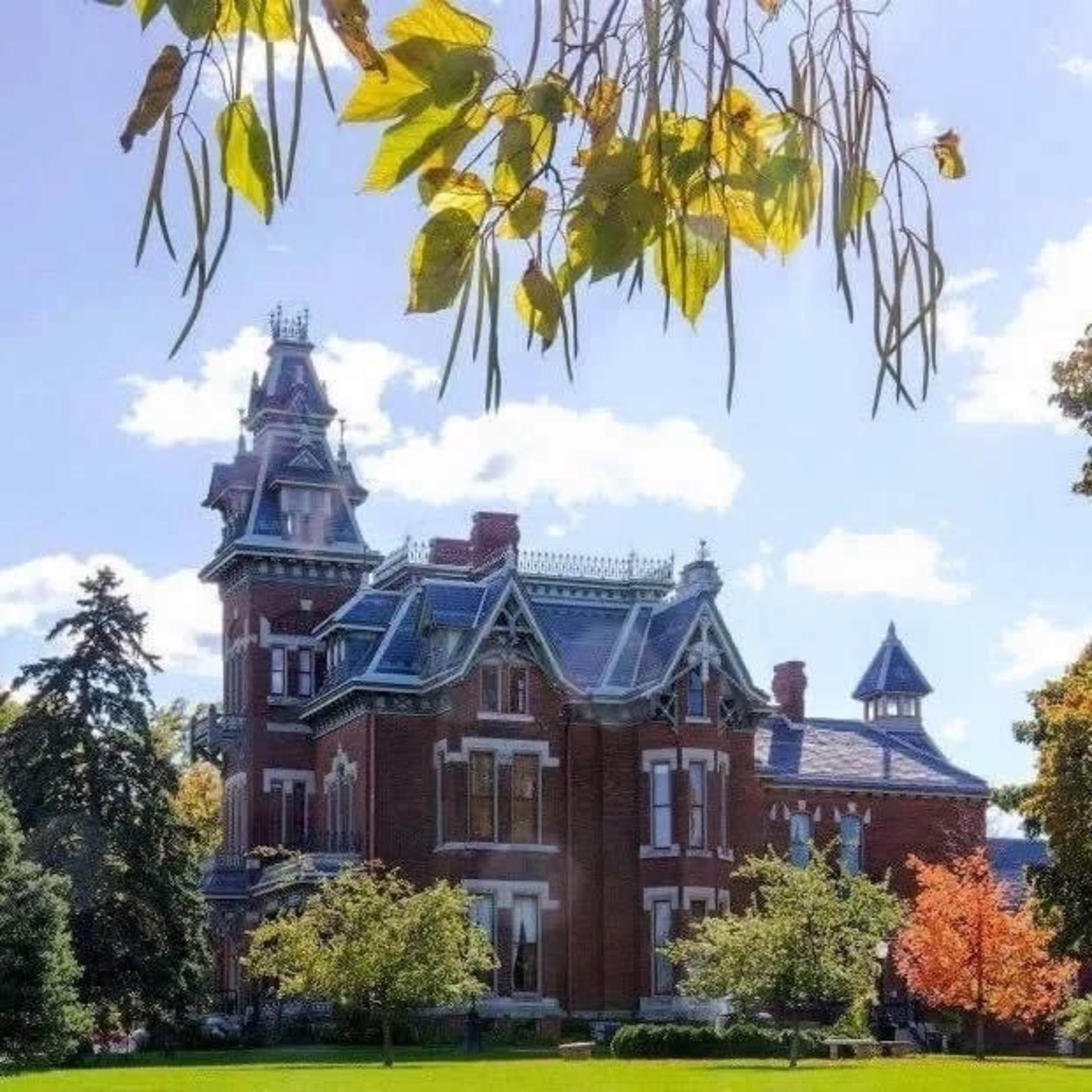 A large Victorian-style mansion with a turret and intricate architectural details is surrounded by manicured lawns and trees.