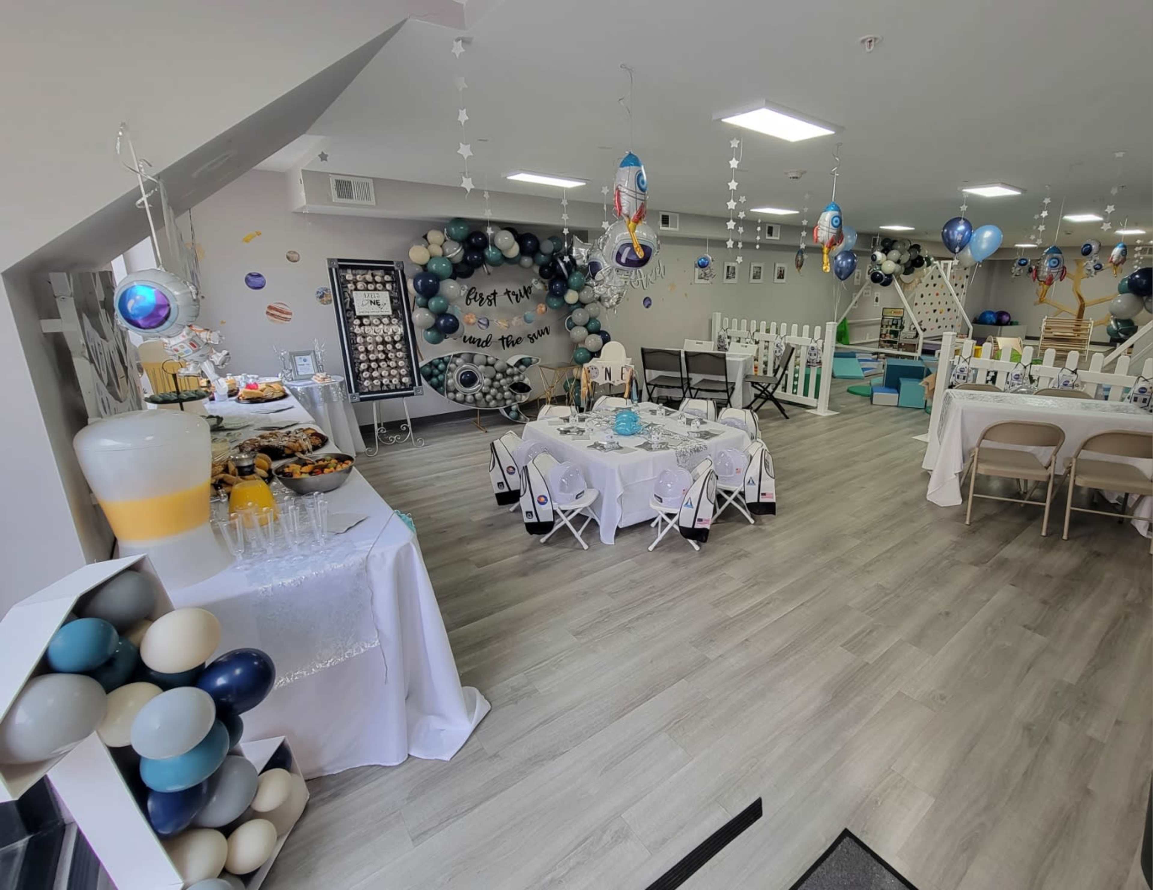 A decorated party venue with tables set for a celebration, featuring a food and drink station and colorful balloons adorning the walls.
