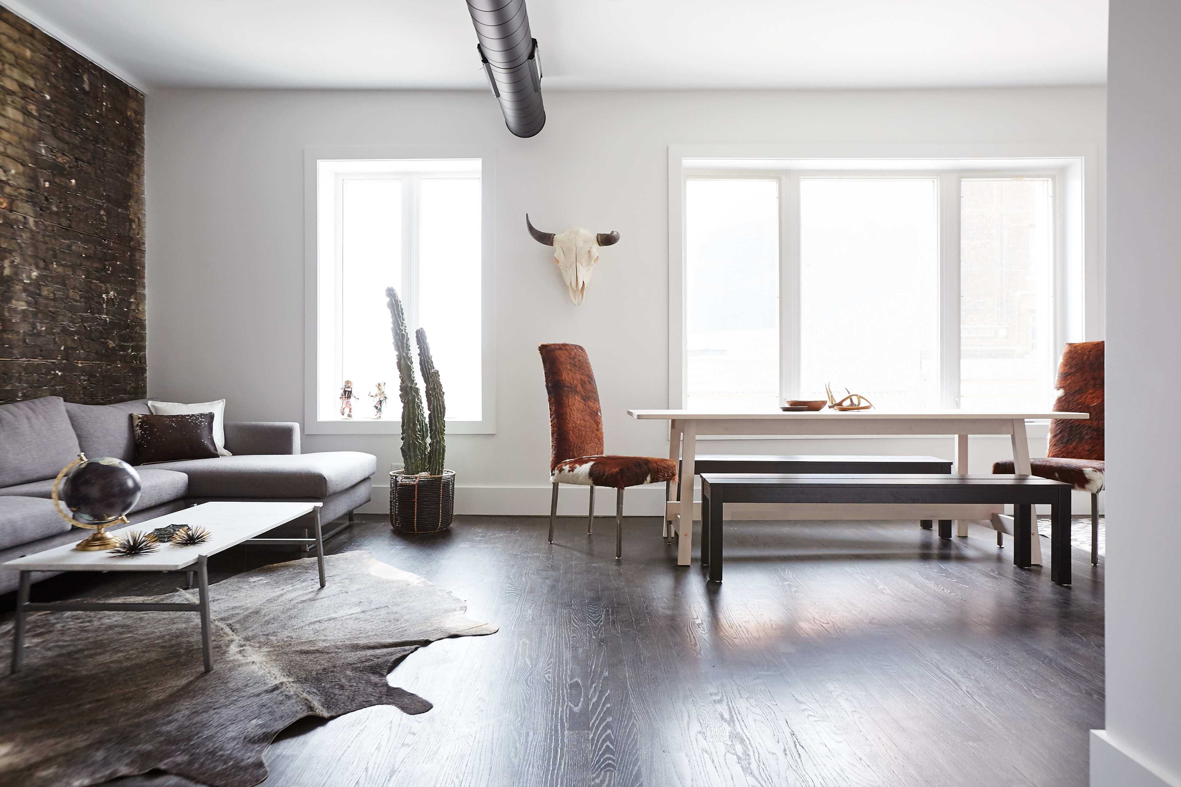 The image features a modern living space with a gray sofa, a dining table surrounded by chairs, and large windows allowing natural light to fill the room.