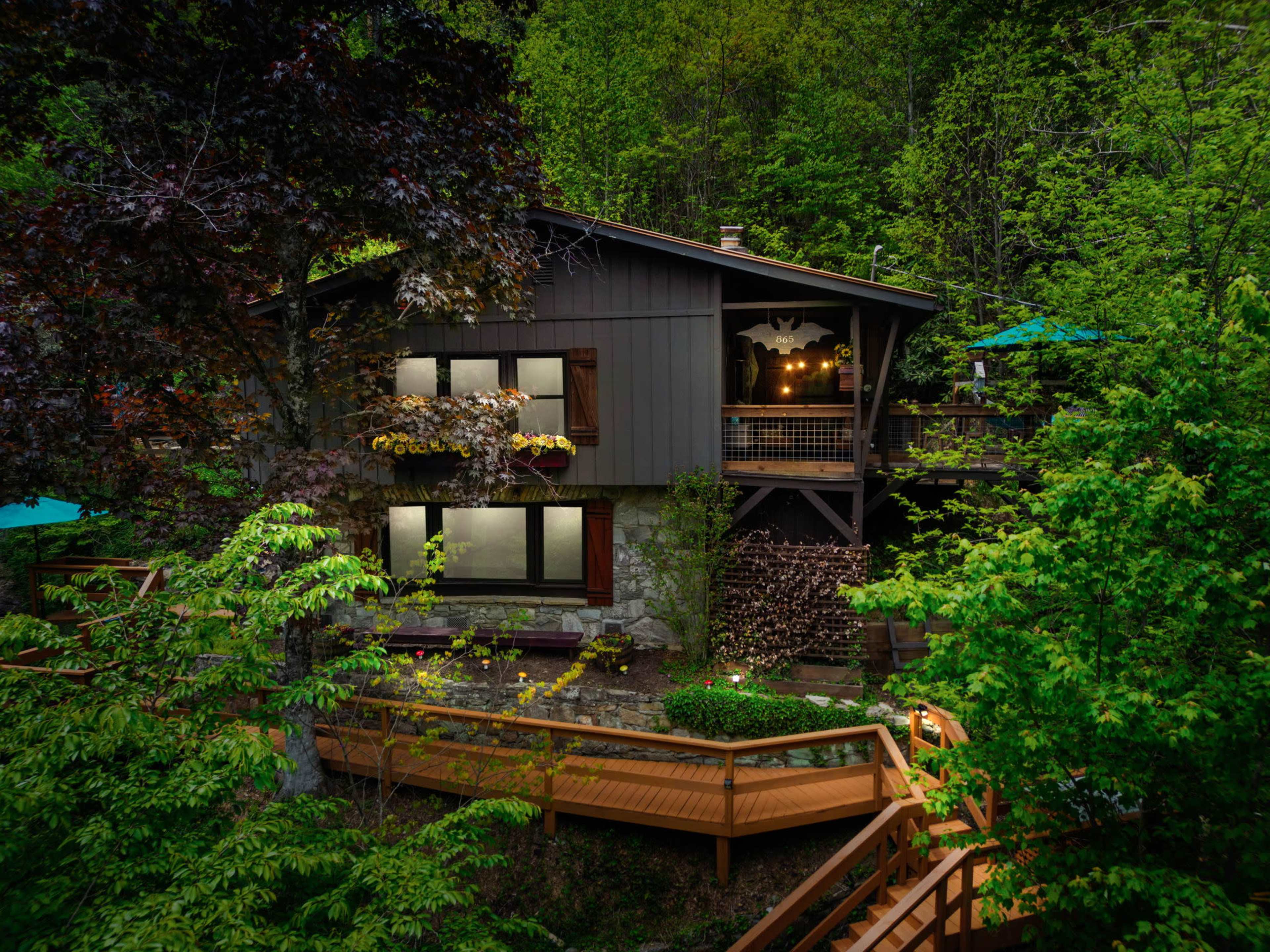 A wooden house with large windows sits nestled among trees, featuring a deck and outdoor seating.