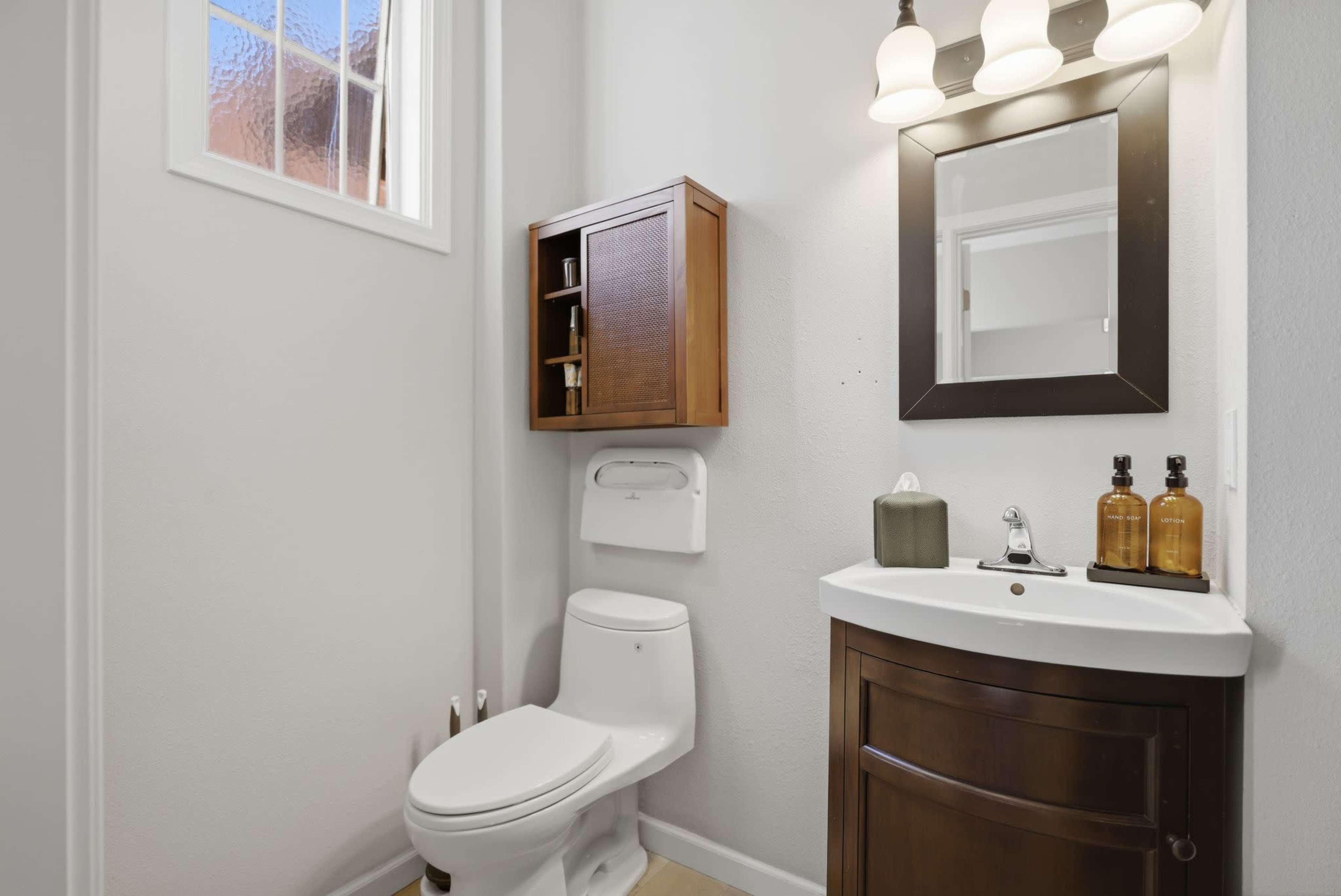 The image shows a small bathroom featuring a toilet, a wooden storage cabinet, a sink with a dark wood vanity, and a mirror with three ceiling-mounted light fixtures.