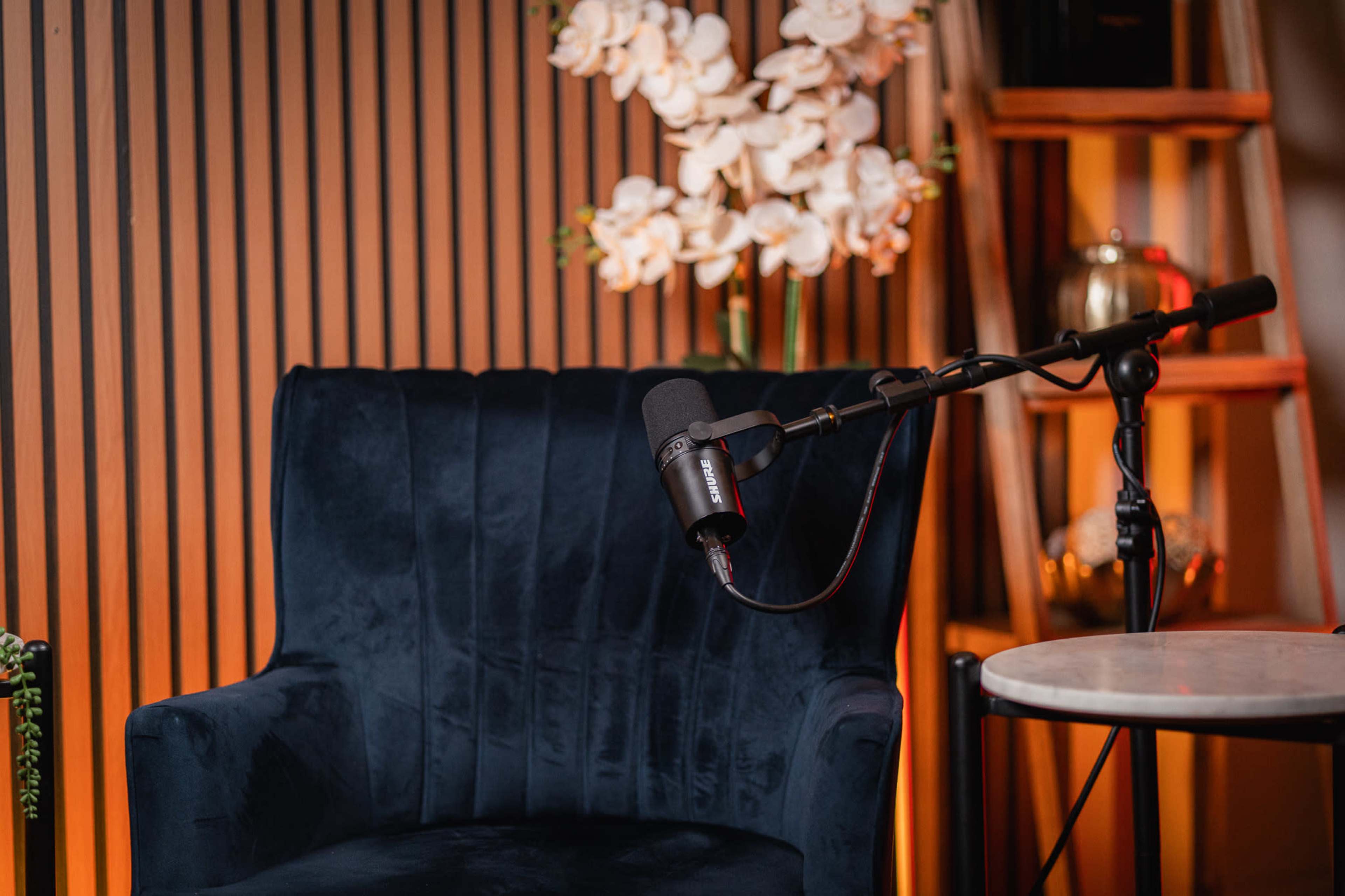The image shows a blue upholstered armchair with a microphone mounted on a stand next to a small round table, against a wooden slatted backdrop and a display of flowers.