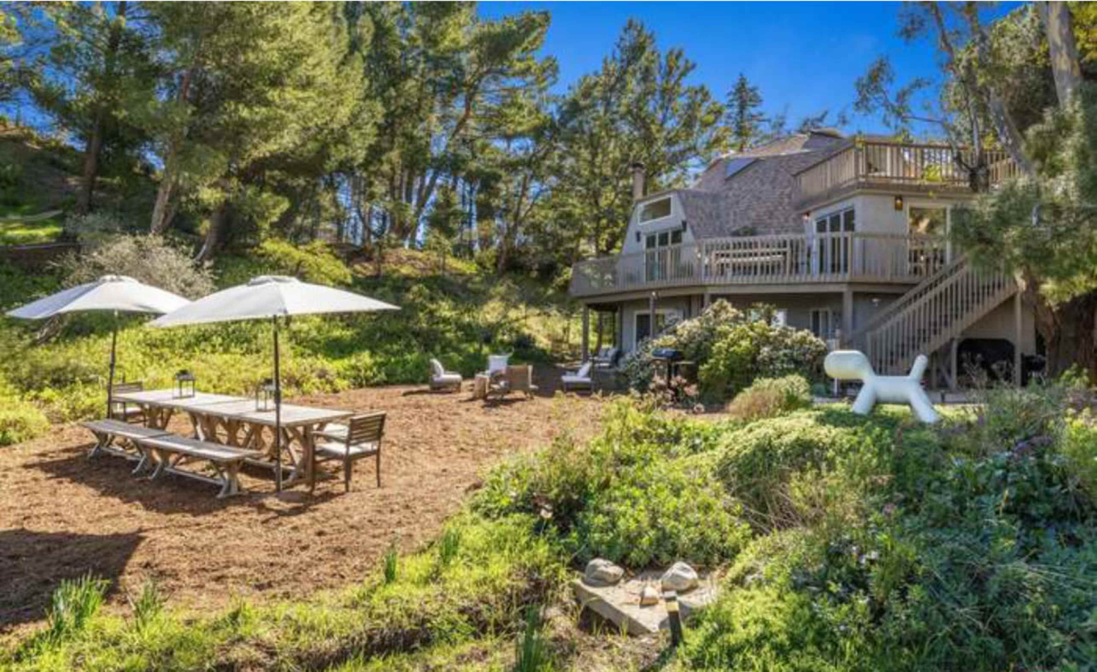 The image shows a spacious backyard with a large wooden dining table under two umbrellas and a modern house in the background surrounded by greenery.