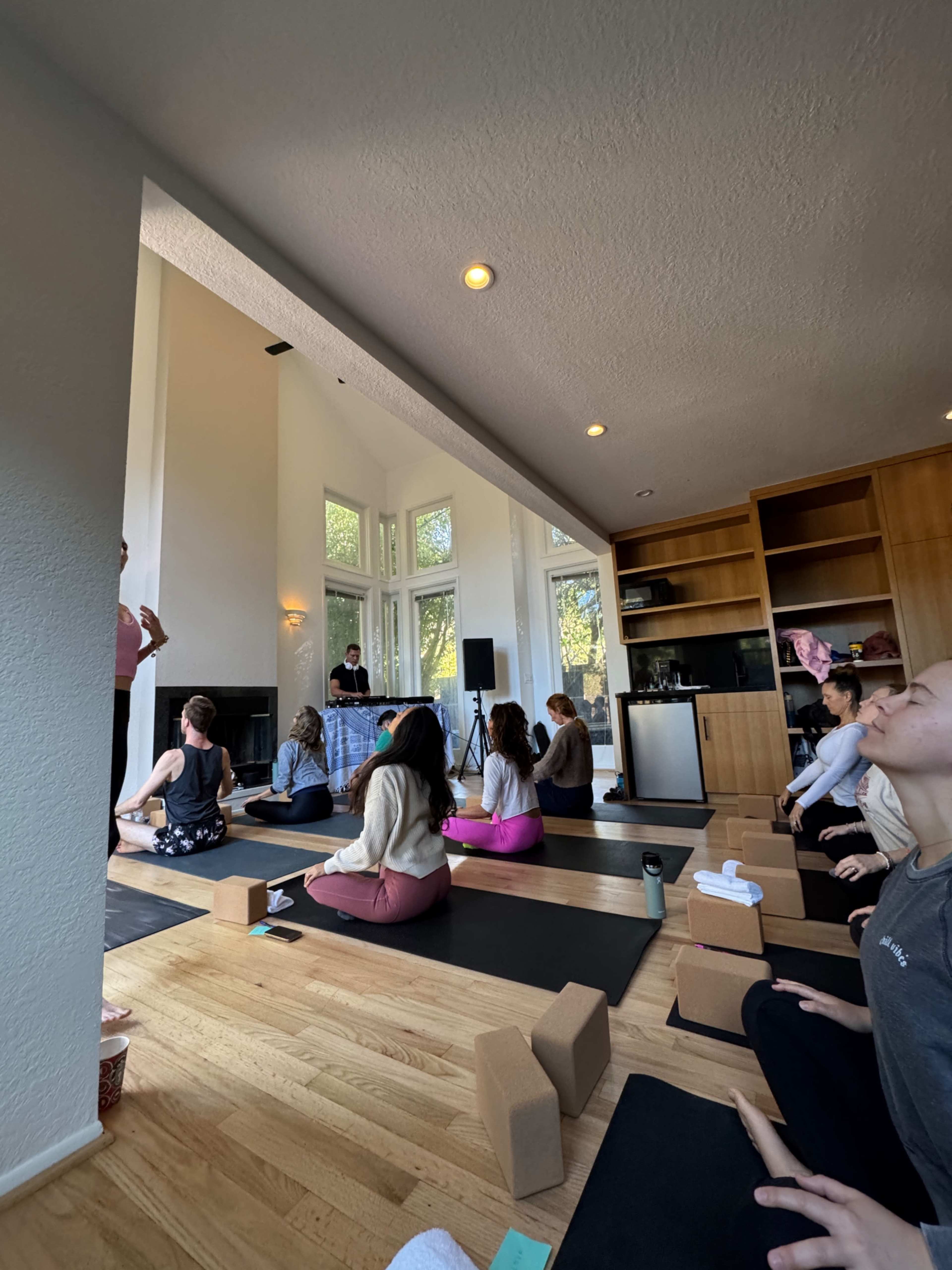 A group of people are seated on yoga mats in a spacious room with large windows and wooden flooring, in a yoga or meditation class.