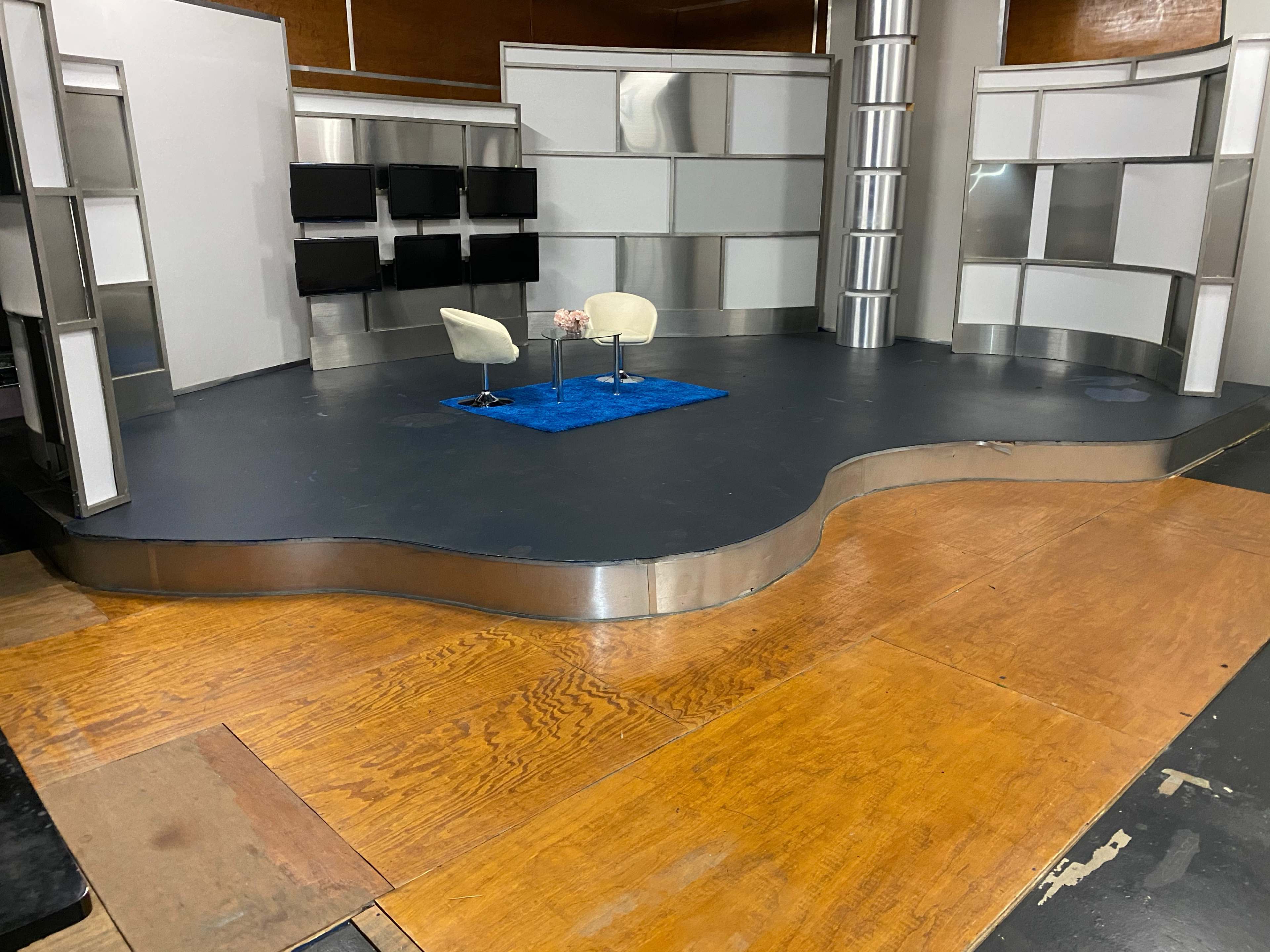 The image shows a television studio set with a curved wooden stage, two white chairs, and a blue carpet, surrounded by gray and silver background panels.