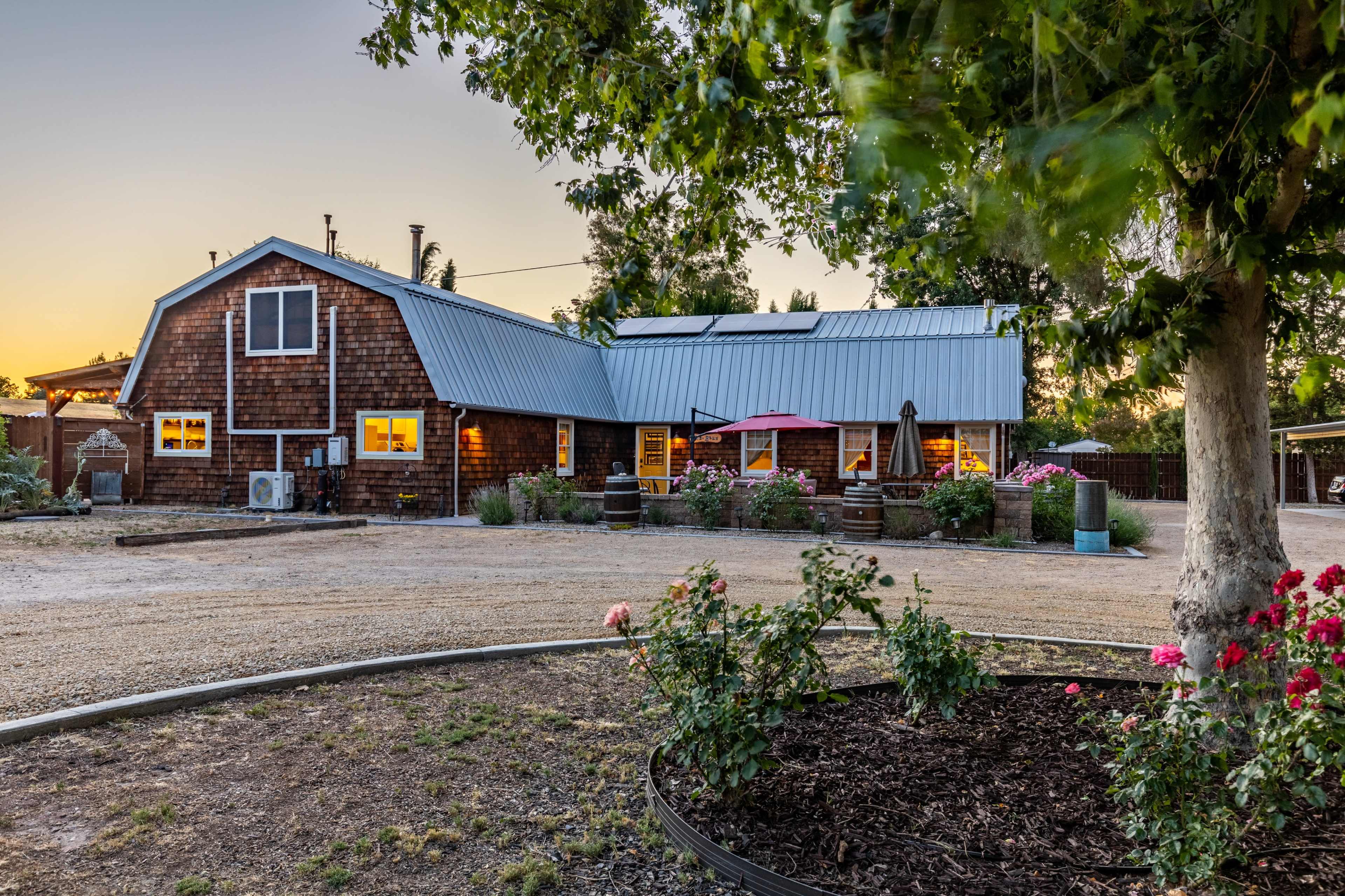 A rustic wooden house with a metal roof, surrounded by a gravel driveway and flower beds at sunset.