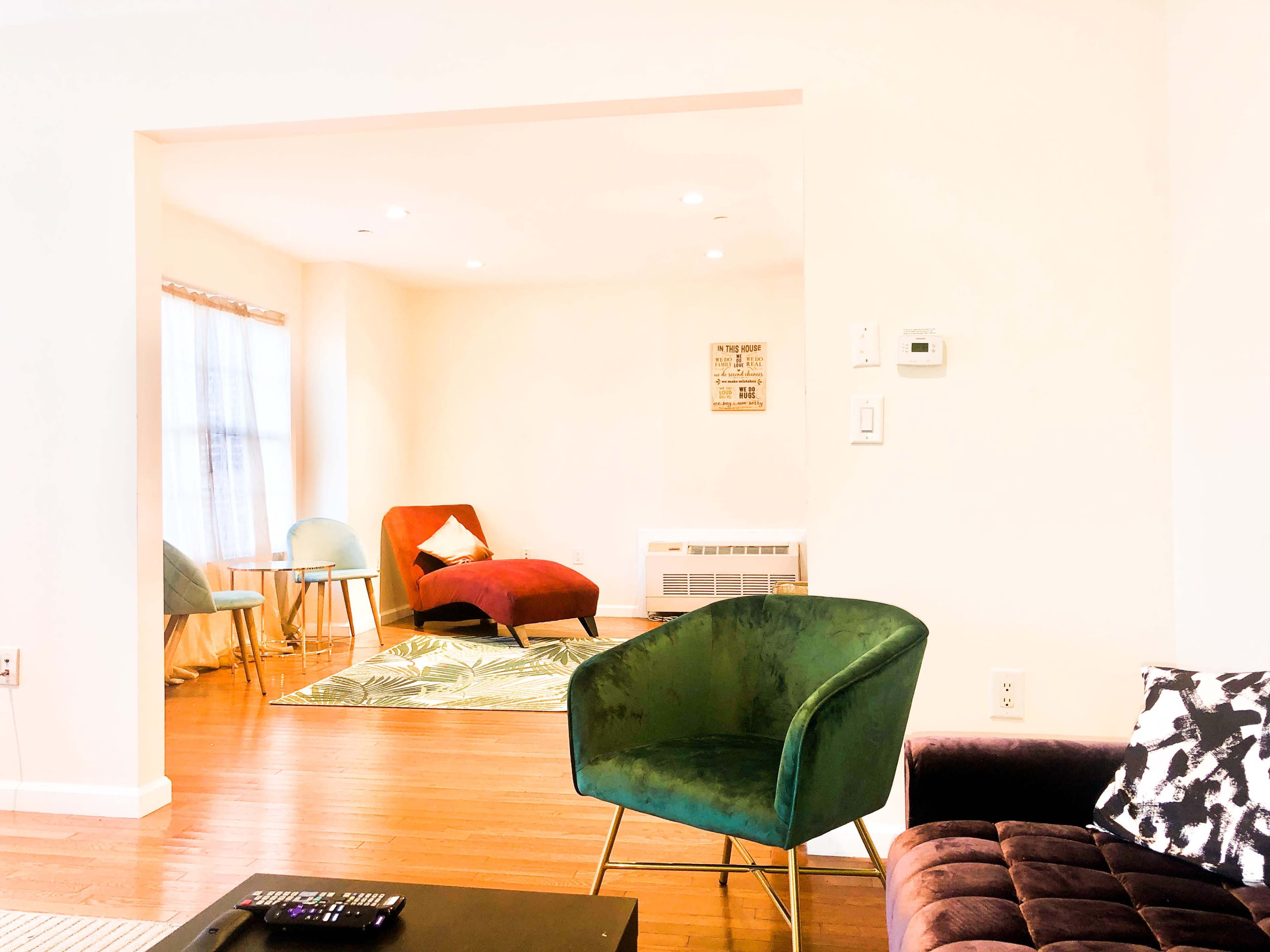 A brightly lit living area features a green chair, a maroon accent chair, and other seating with a patterned rug on wooden flooring.