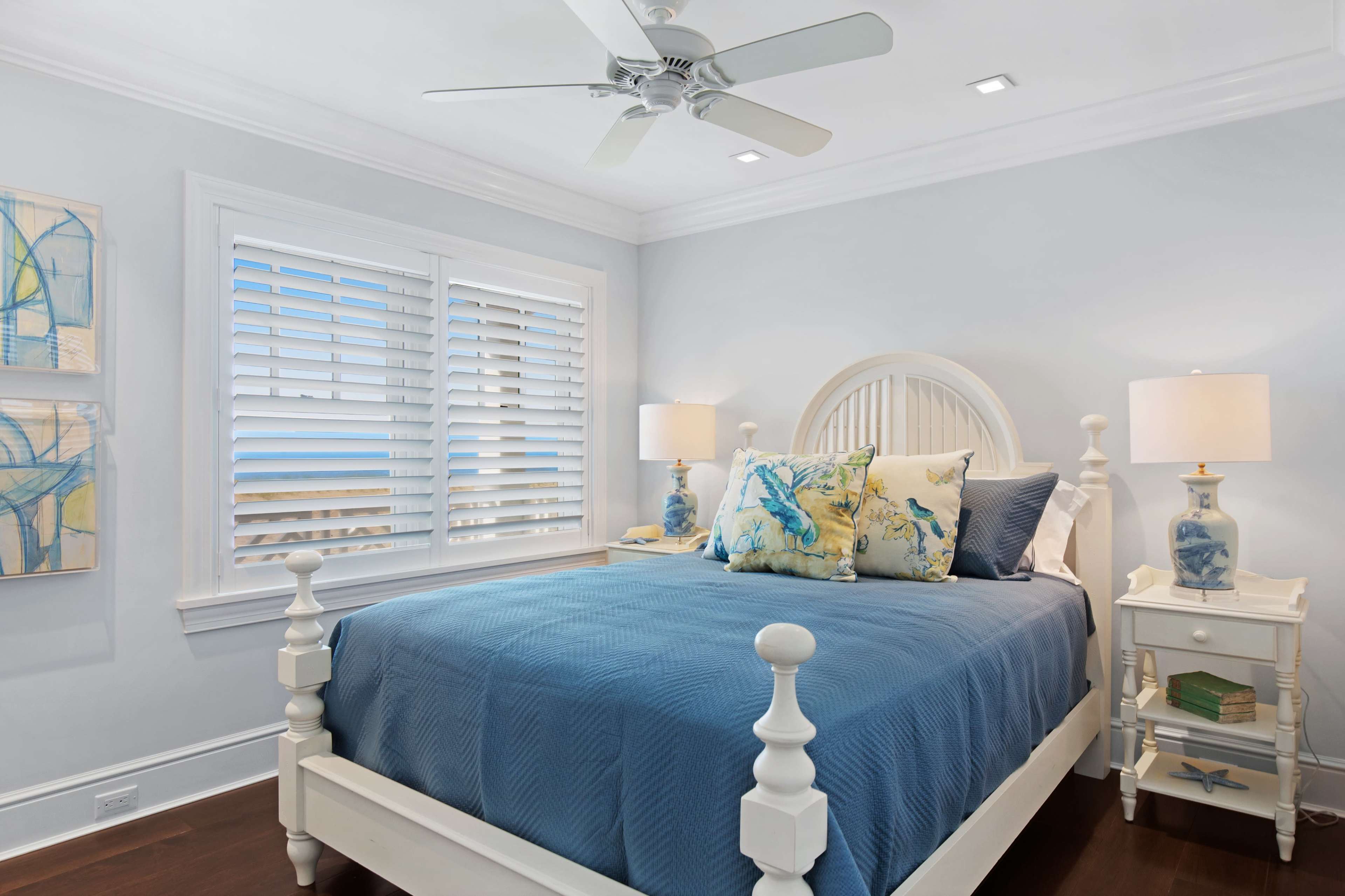 A light-colored bedroom features a white bed with a blue quilt and decorative pillows, alongside a nightstand with a lamp and artwork on the wall.