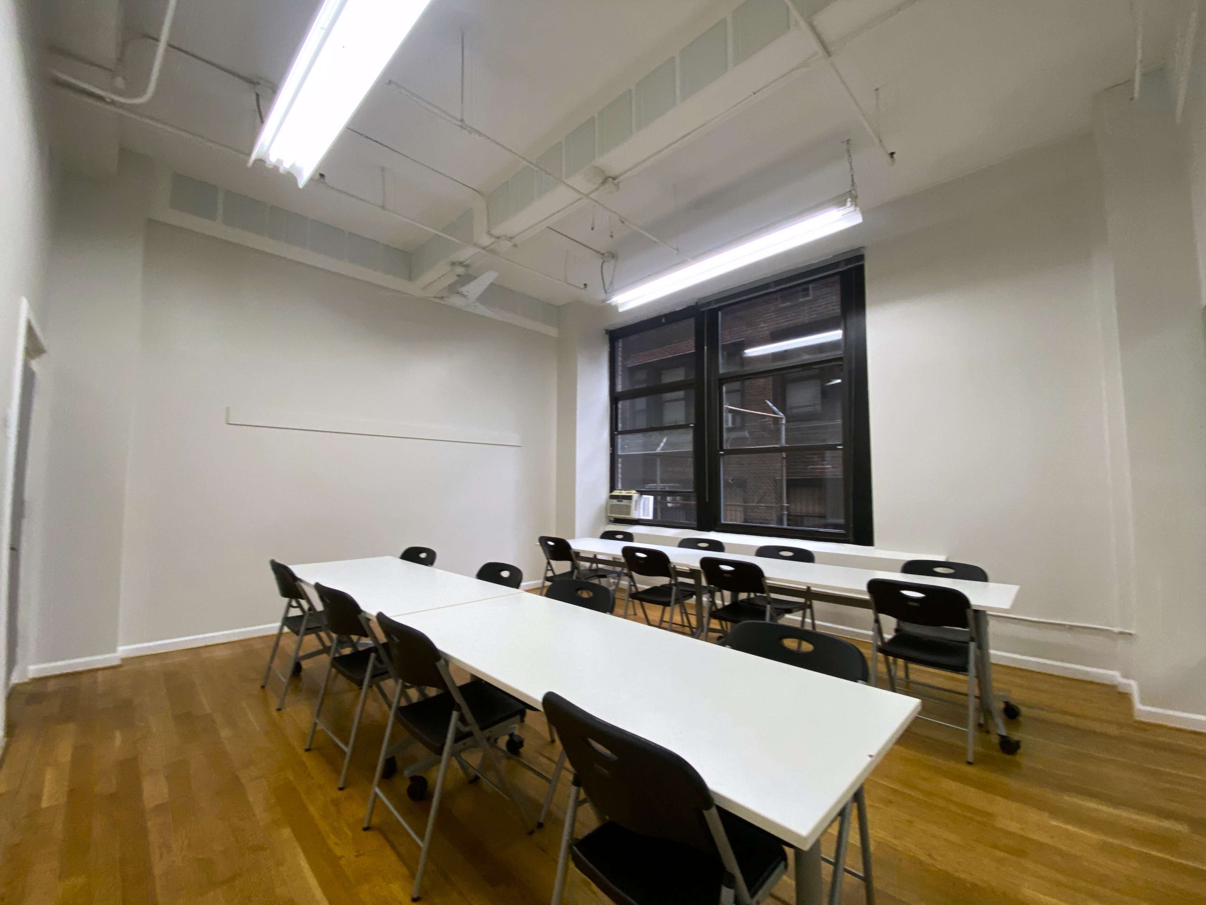 A simple classroom features a long table with chairs arranged around it and a large window letting in natural light.