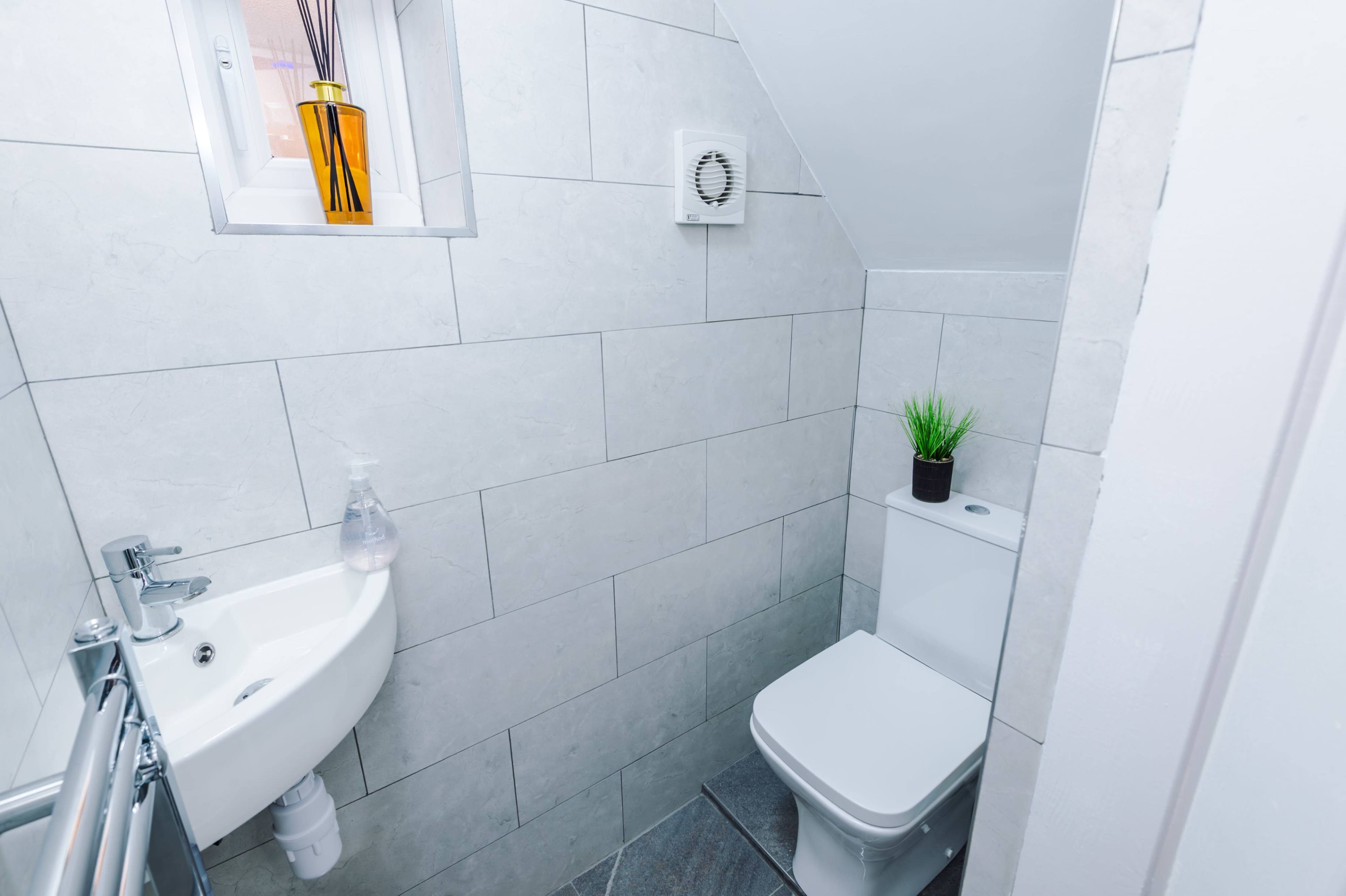 The image shows a small bathroom with gray tiled walls, a corner sink, and a toilet.