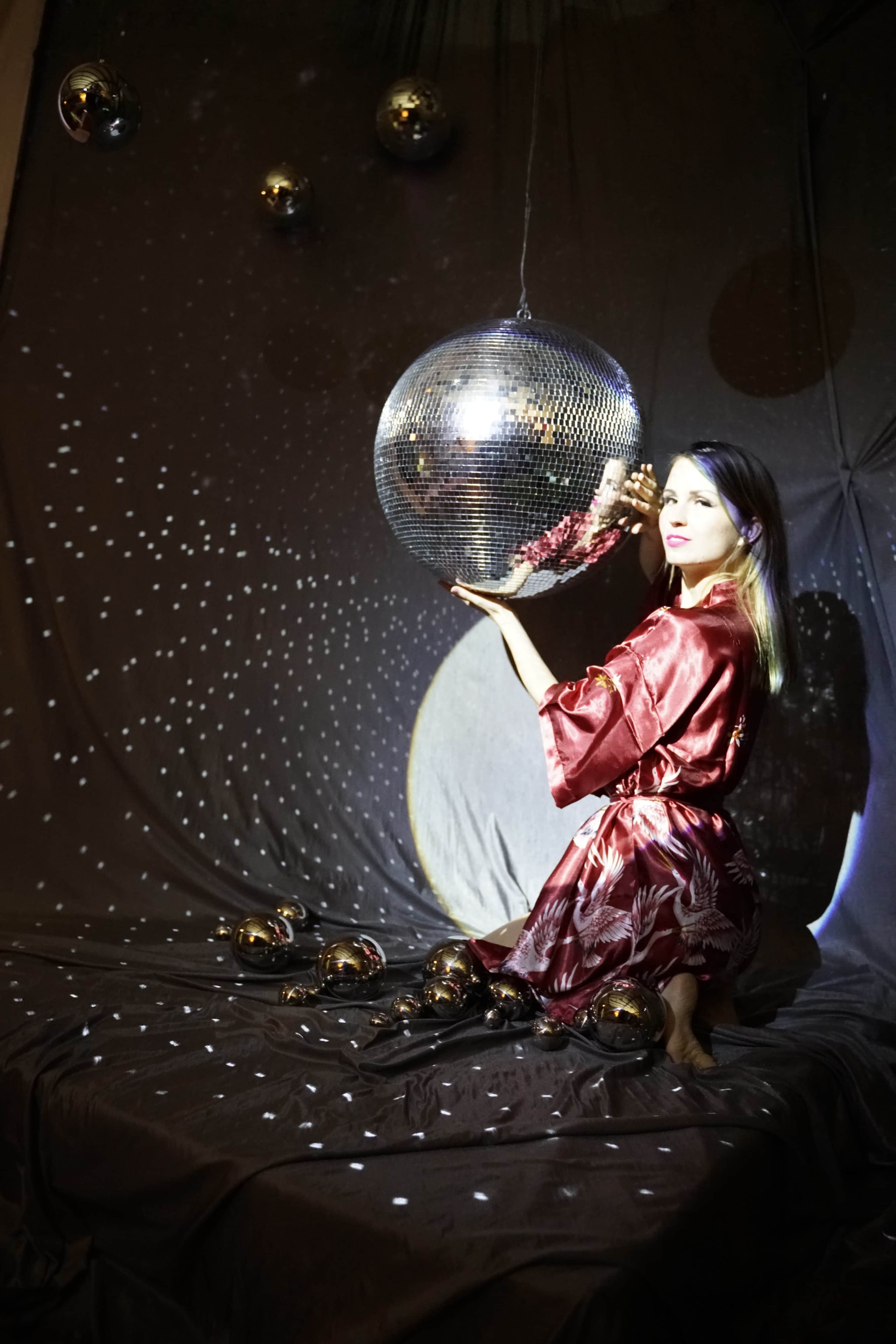 A person in a red robe holds a disco ball amidst a backdrop of textured fabric and scattered light reflections.