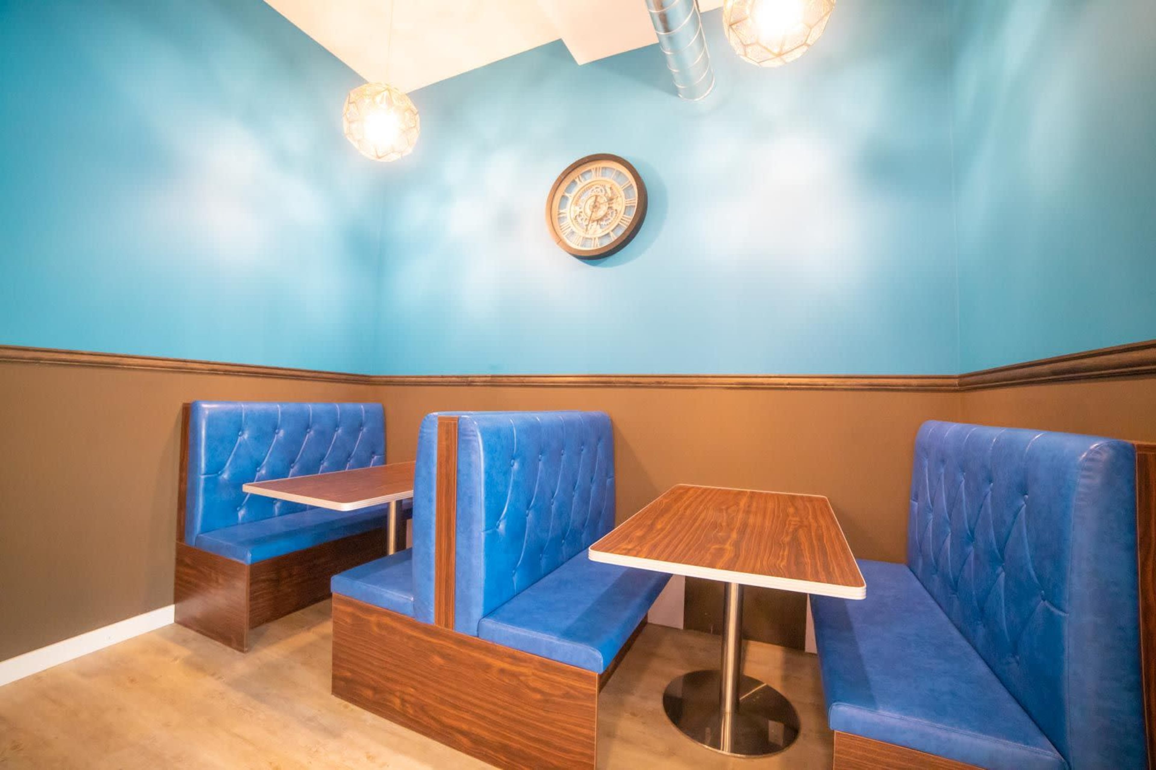 A small dining area with two blue upholstered booths and wooden tables against a turquoise and brown wall, featuring a round clock on the wall.