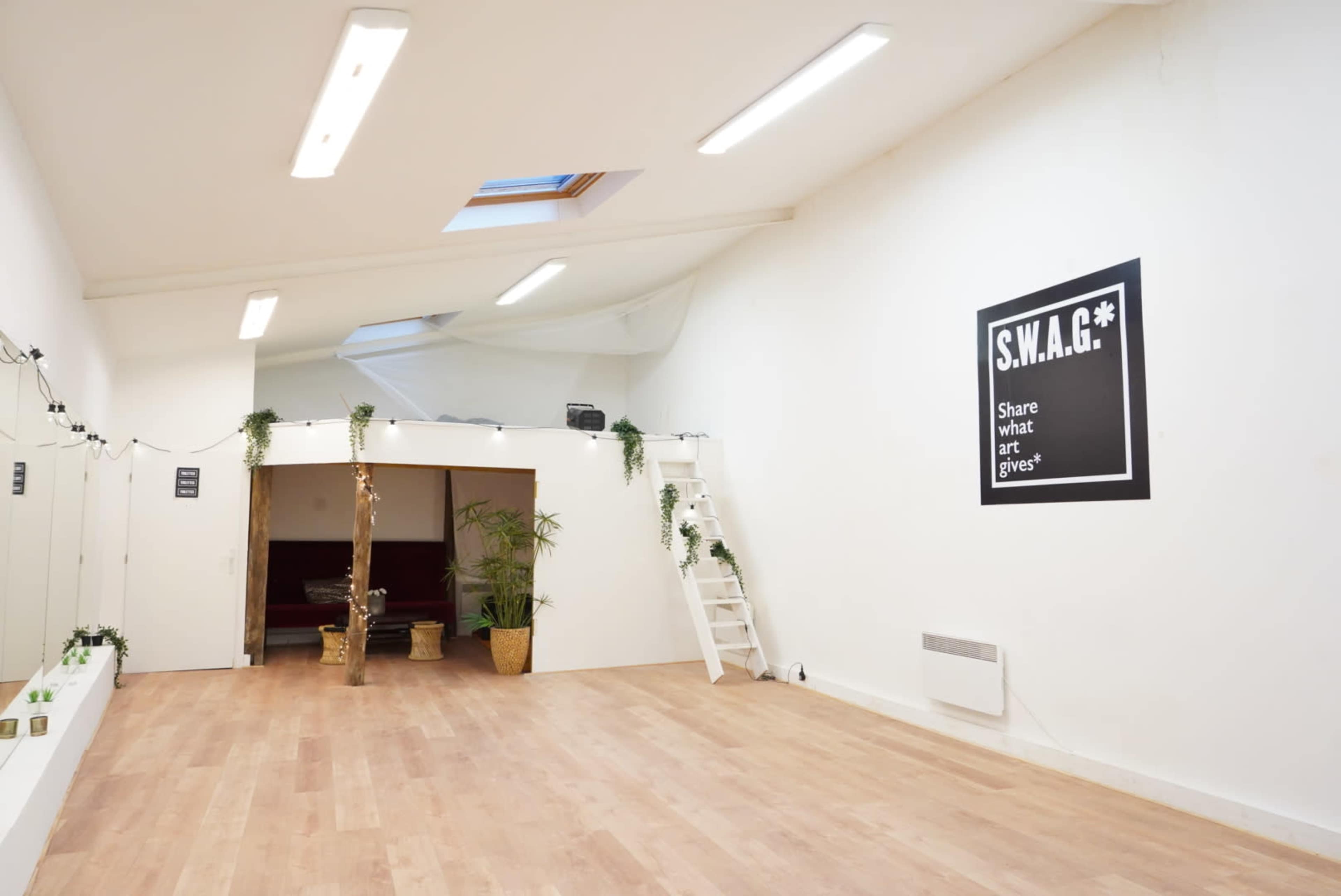 A spacious, well-lit room with wooden flooring, a high ceiling, and minimalist decor, including a wall with the text "S.W.A.G.*" and potted plants.