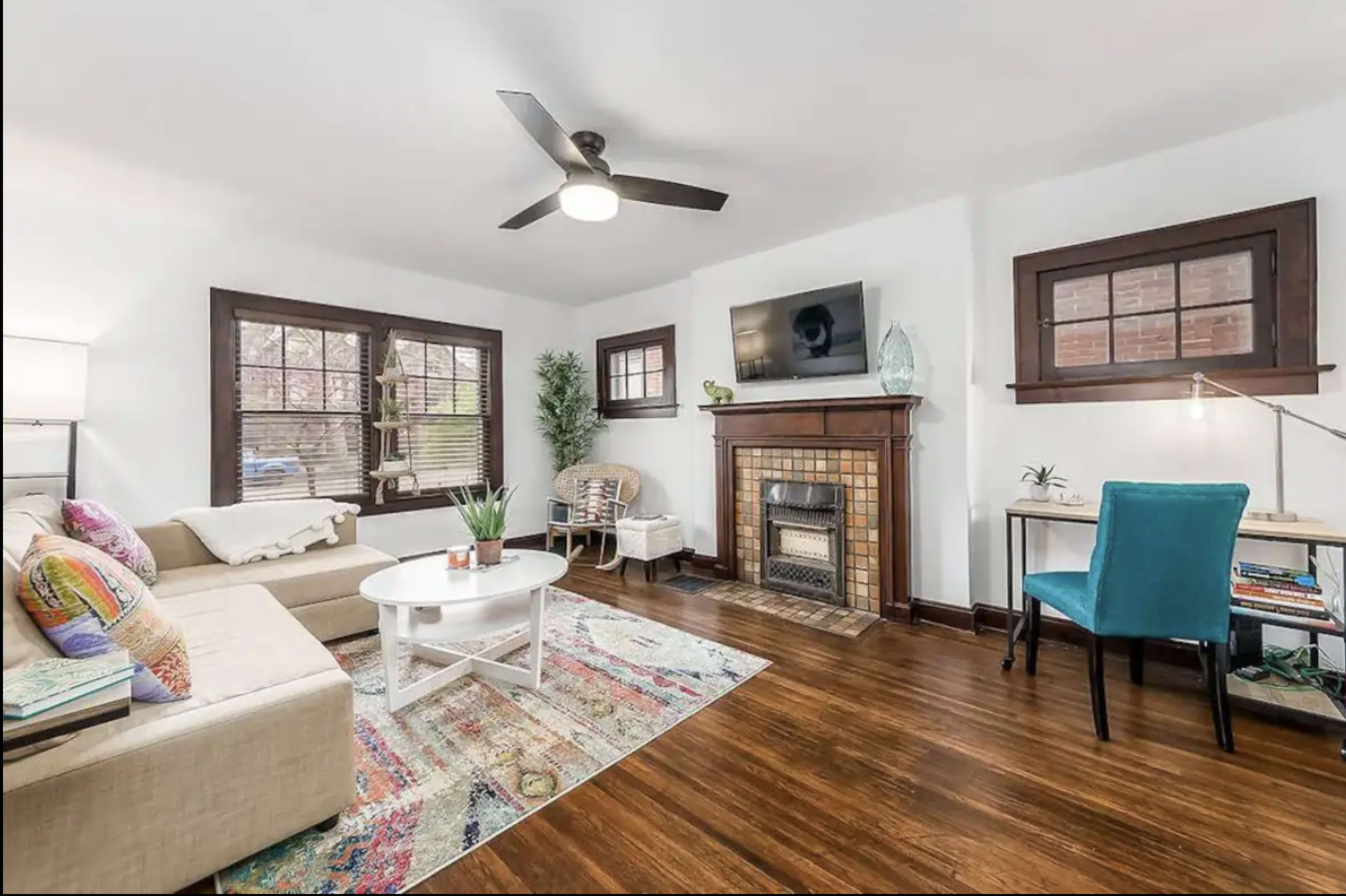 A cozy living room featuring a beige sectional sofa, a round coffee table, a fireplace with a TV mounted above it, and a small workspace with a blue chair.