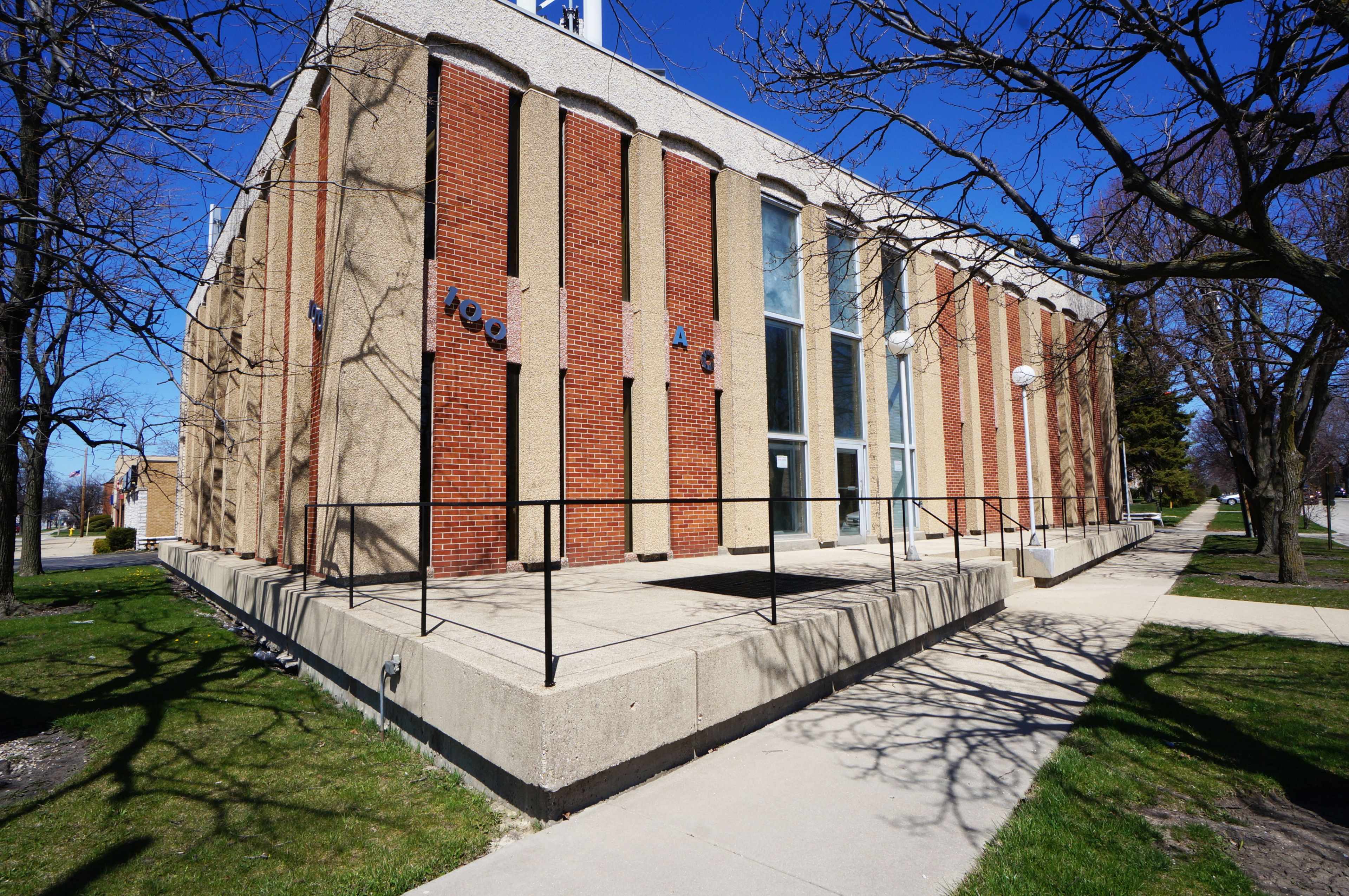 A modern brick building with large windows and a concrete walkway, surrounded by grass and bare trees in a clear blue sky.