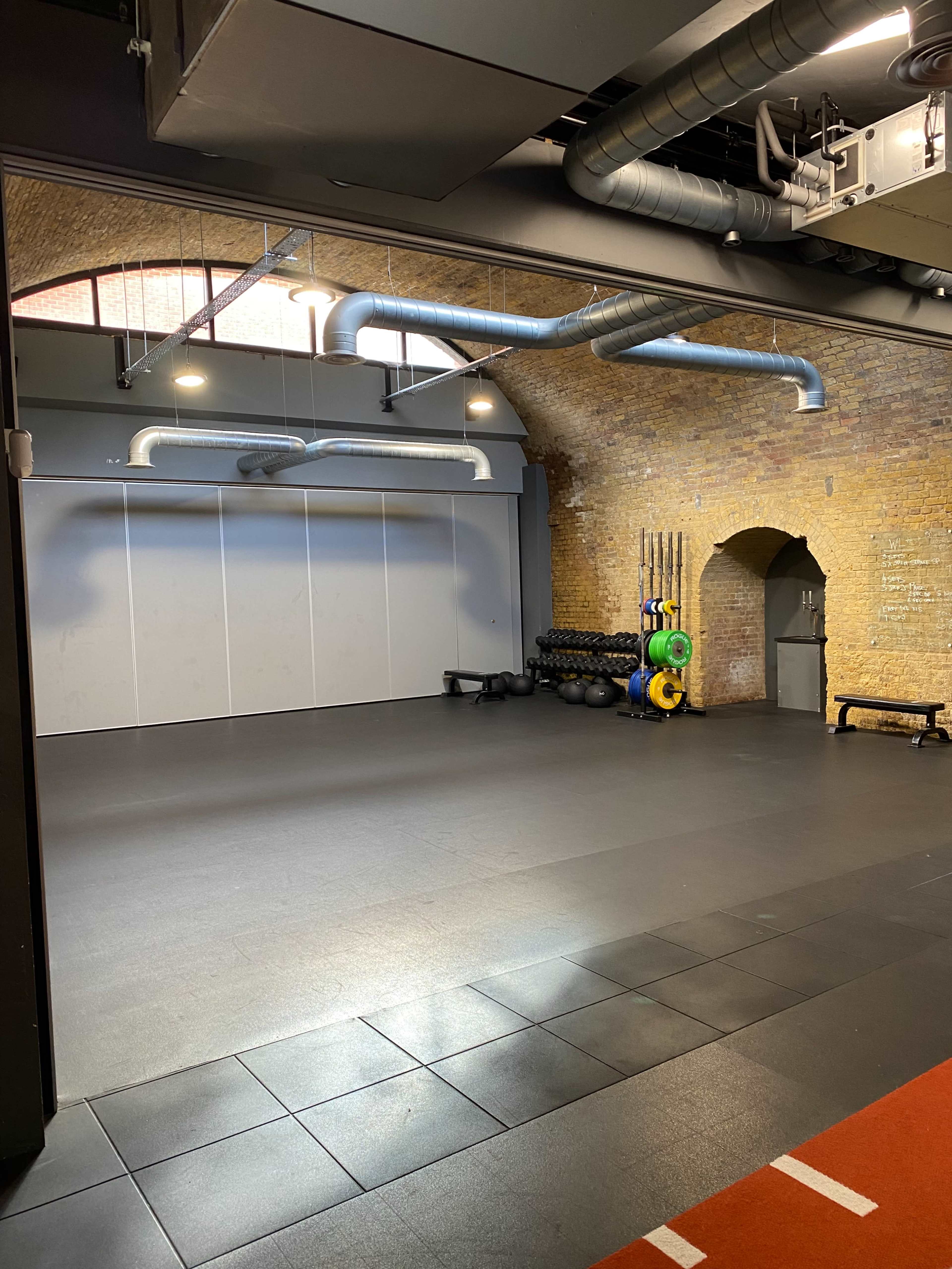 An empty gym space with a rubber floor, exposed brick walls, and various weights stored against one side.