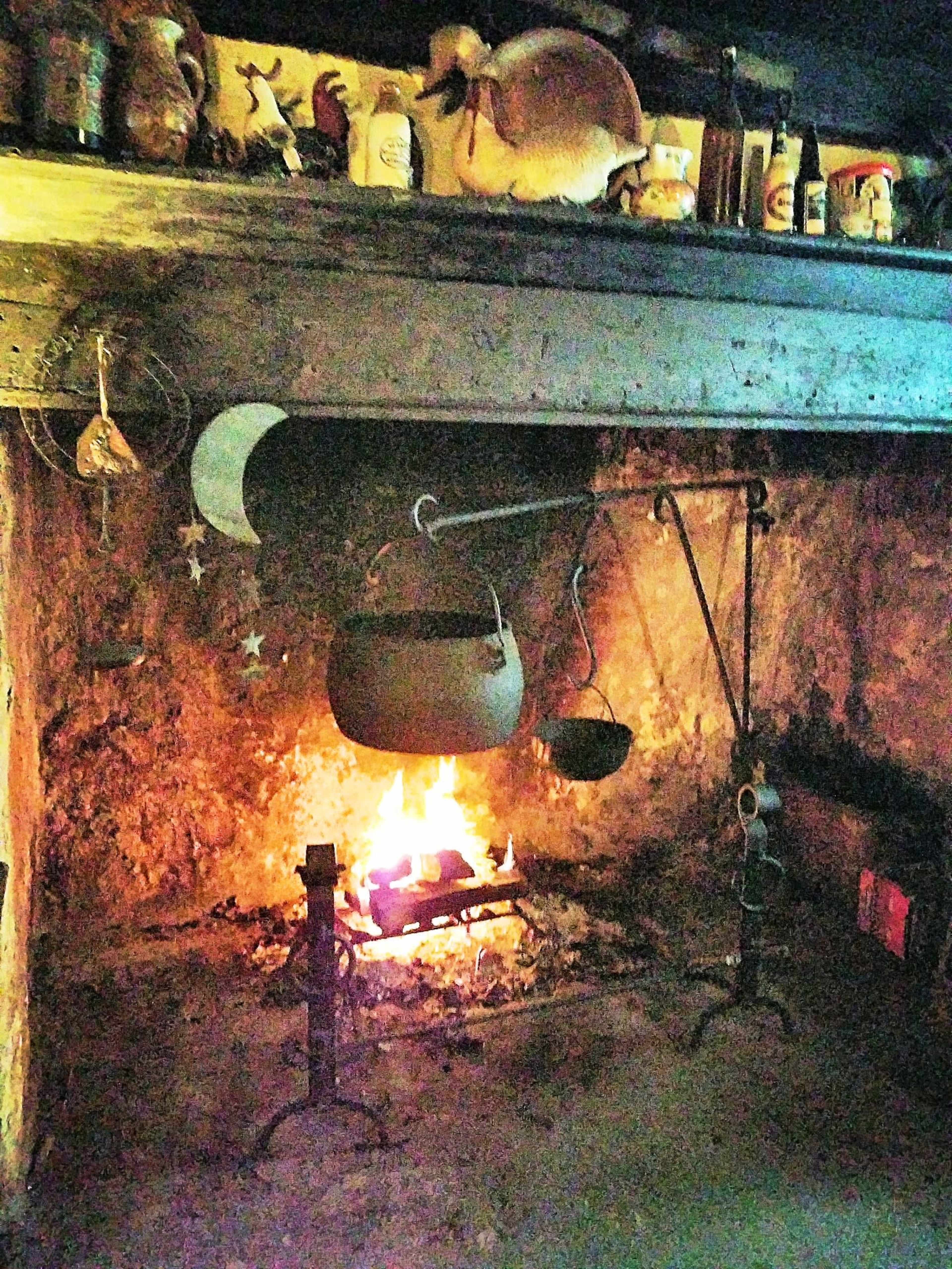 An old stone fireplace with a fire burning, a hanging cauldron on a wrought iron stand, and decorative elements on the mantel.