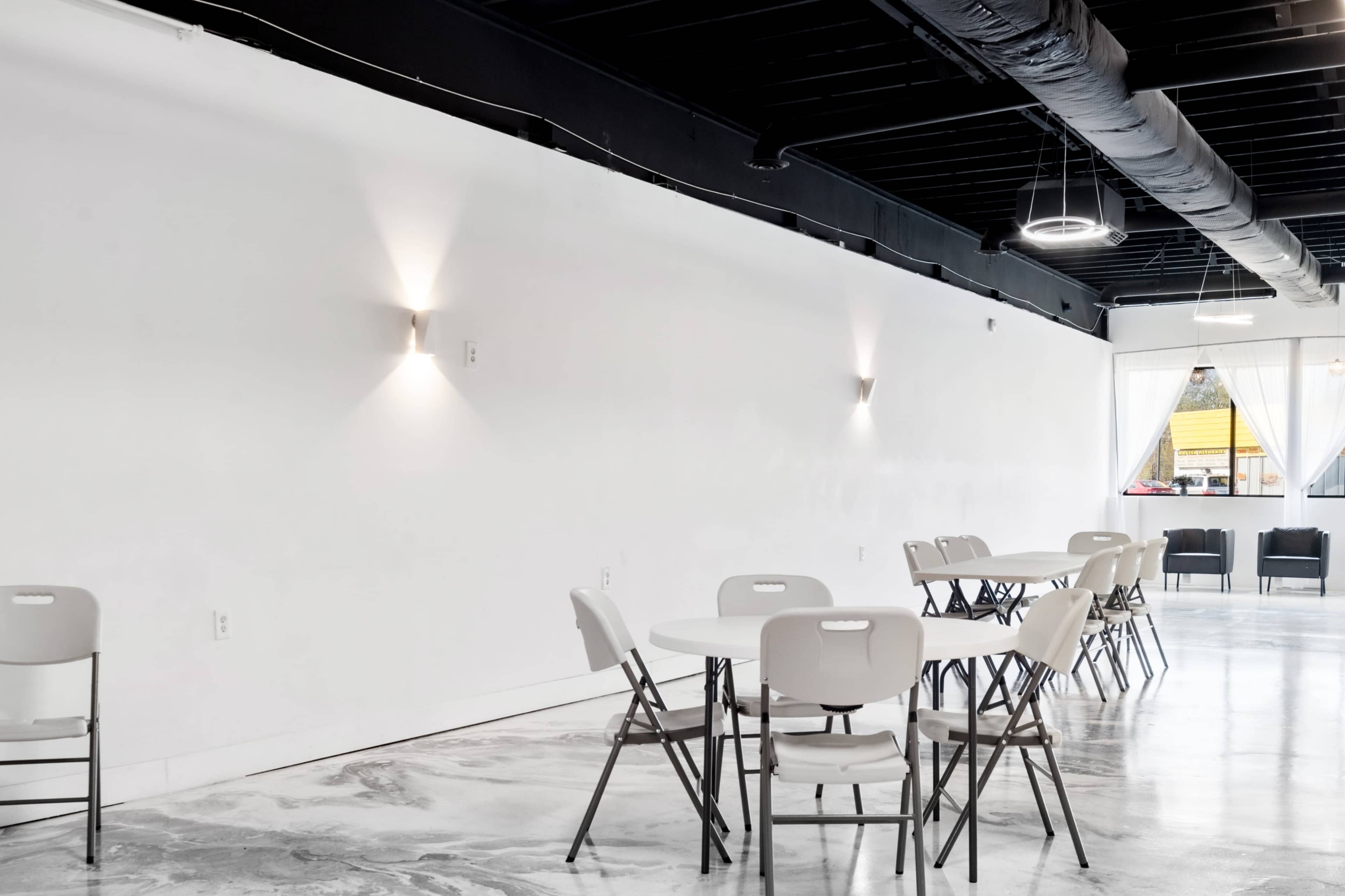 The image shows a modern, minimalist event space with white walls, circular tables, and folding chairs arranged on a polished floor.