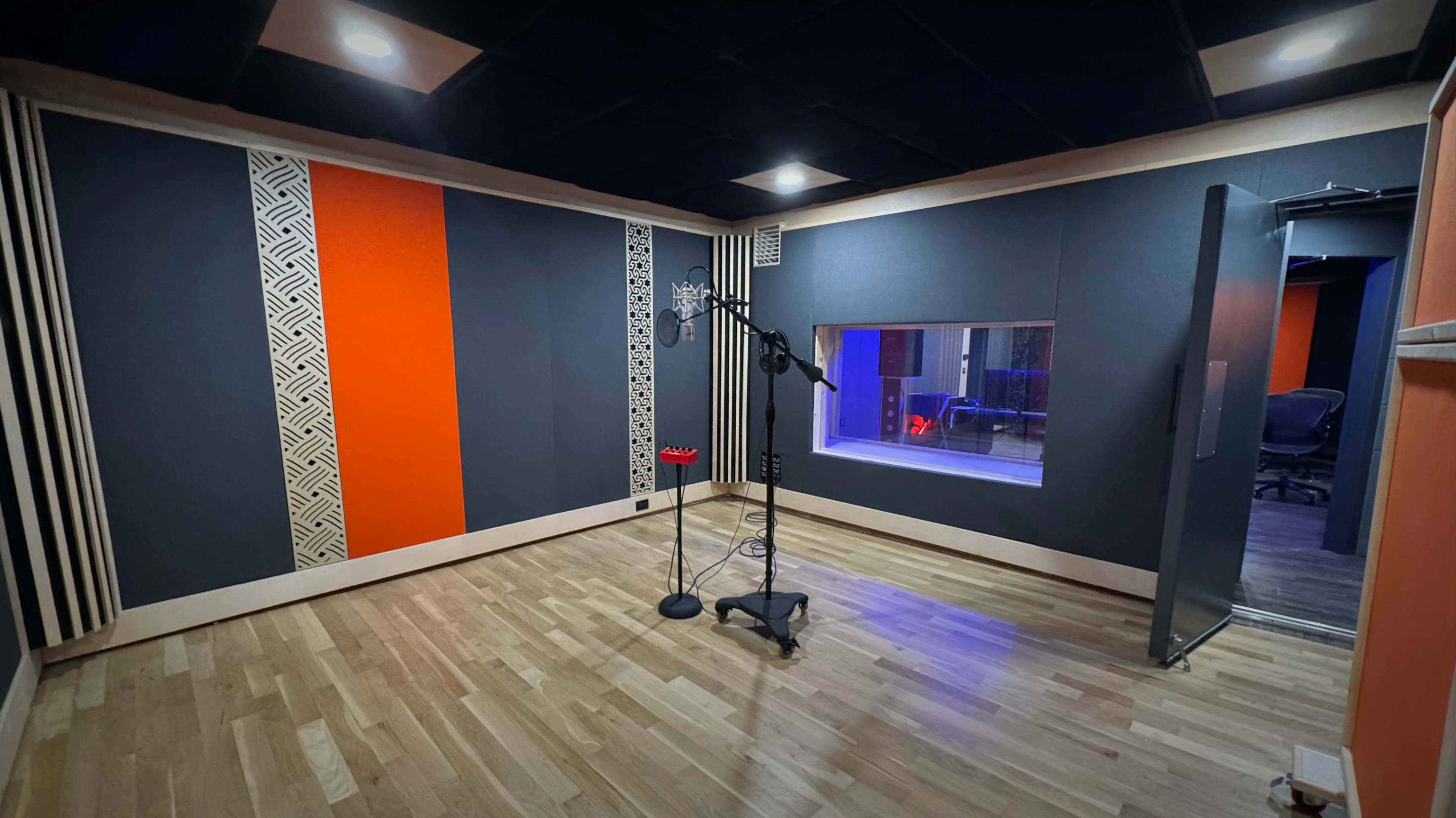 The image shows a soundproof recording studio featuring a microphone stand, decorative wall panels in orange and gray, and a large window overlooking another room.