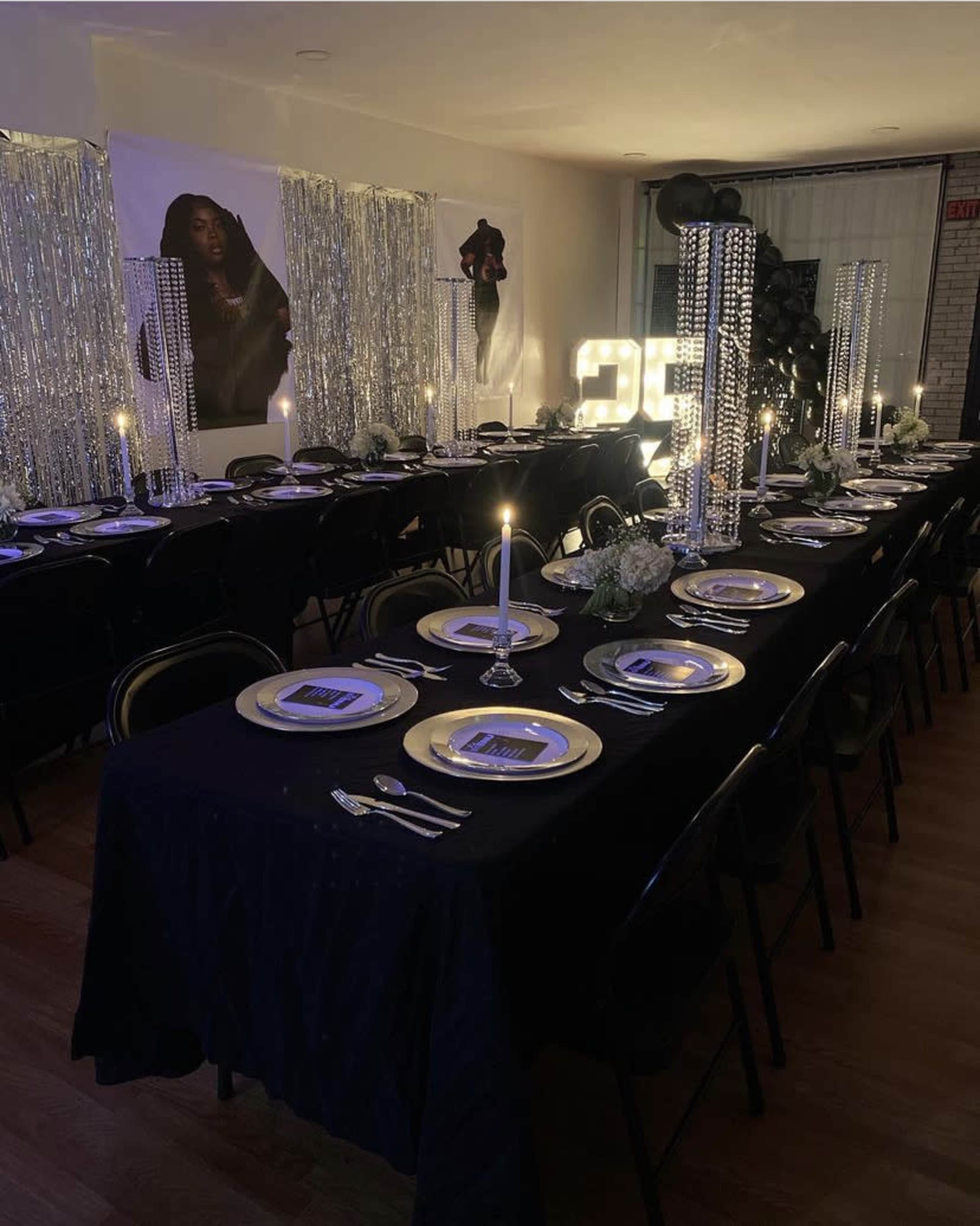 A dining area is set for a formal event, featuring a long black table with elegant place settings, candles, decorative centerpieces, and illuminated numbers.