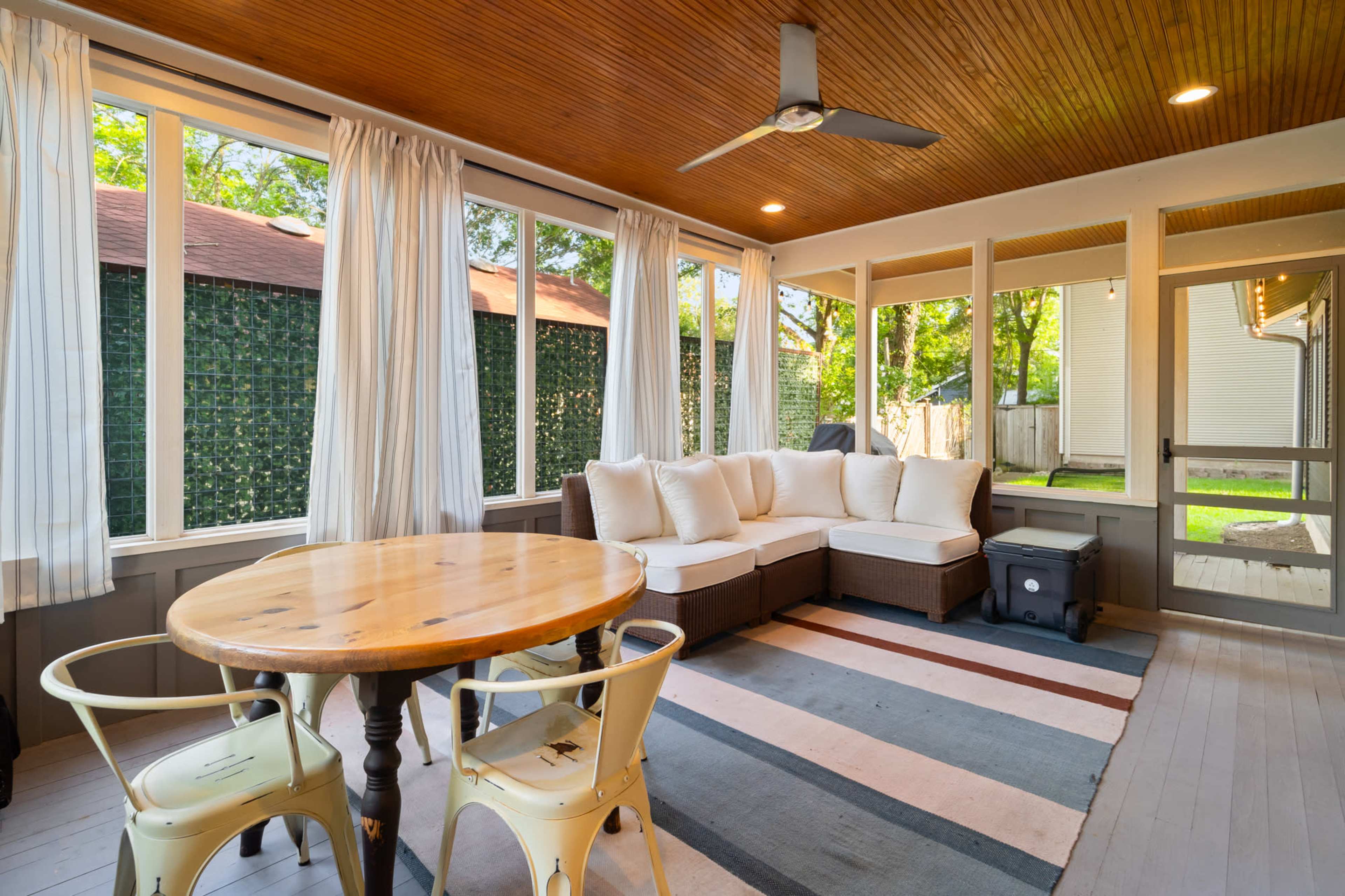 The image shows a sunlit porch with a cozy seating area, a small wooden table, and large windows draped with light curtains.