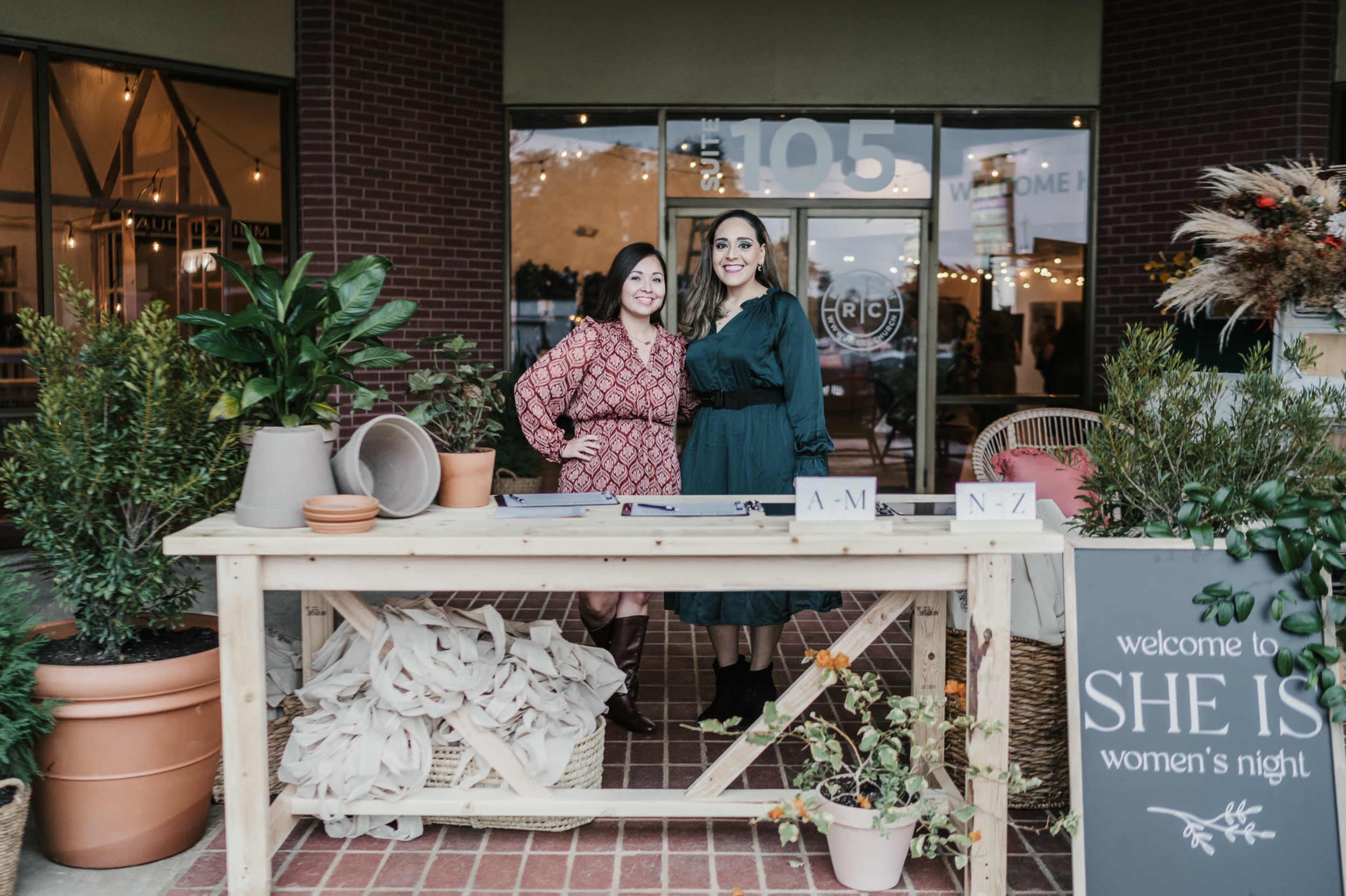 Two women stand behind a wooden table adorned with various items, in front of a storefront with welcoming decorations for an event titled "She Is: Women's Night."