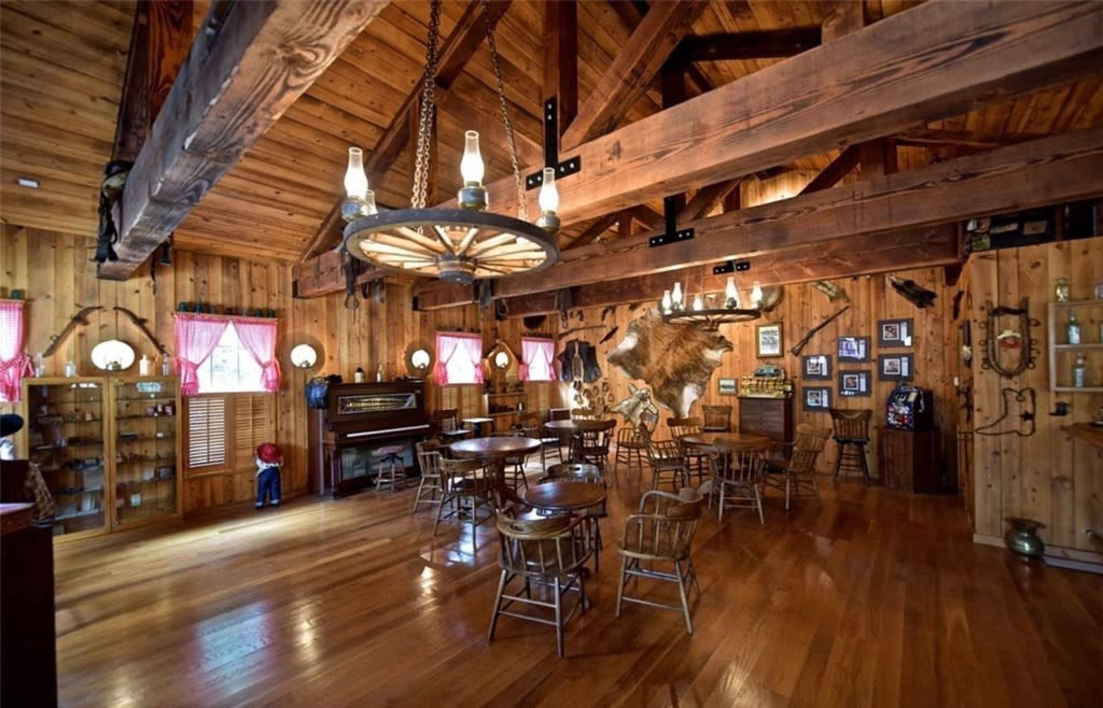 A wooden interior of a rustic dining or gathering space, featuring exposed beams, several round tables, and decorations including a mounted animal head and a piano.