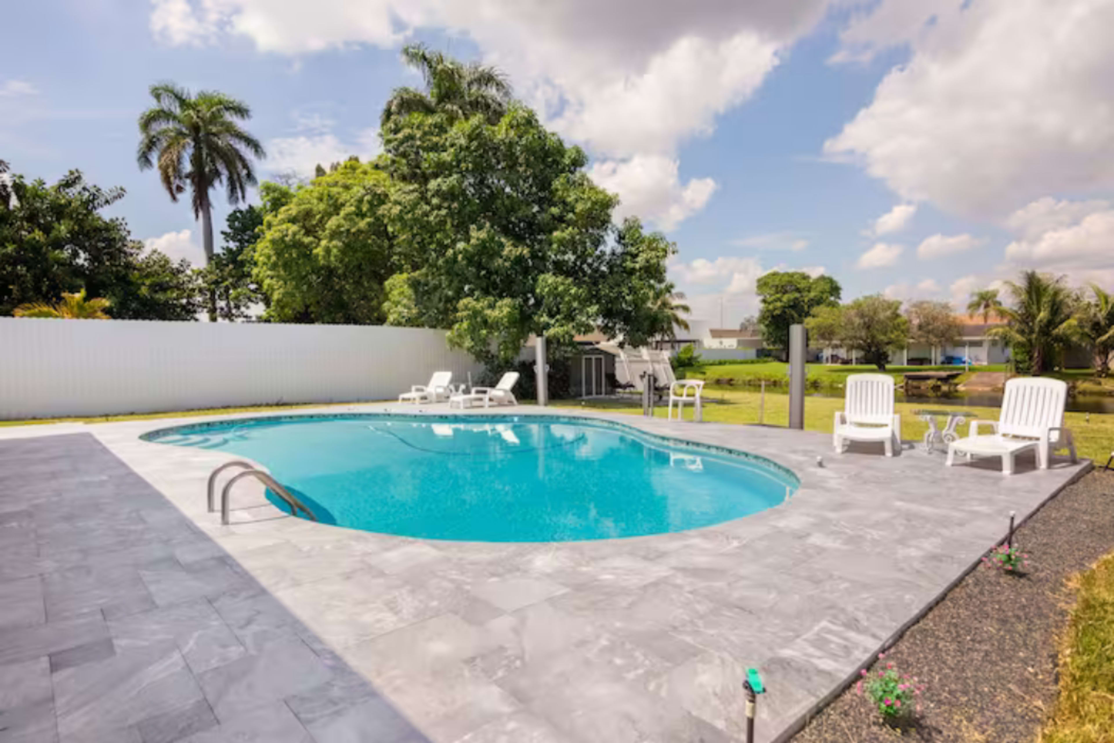 A rectangular swimming pool is surrounded by a patio with lounge chairs, set against a backdrop of greenery and blue skies.