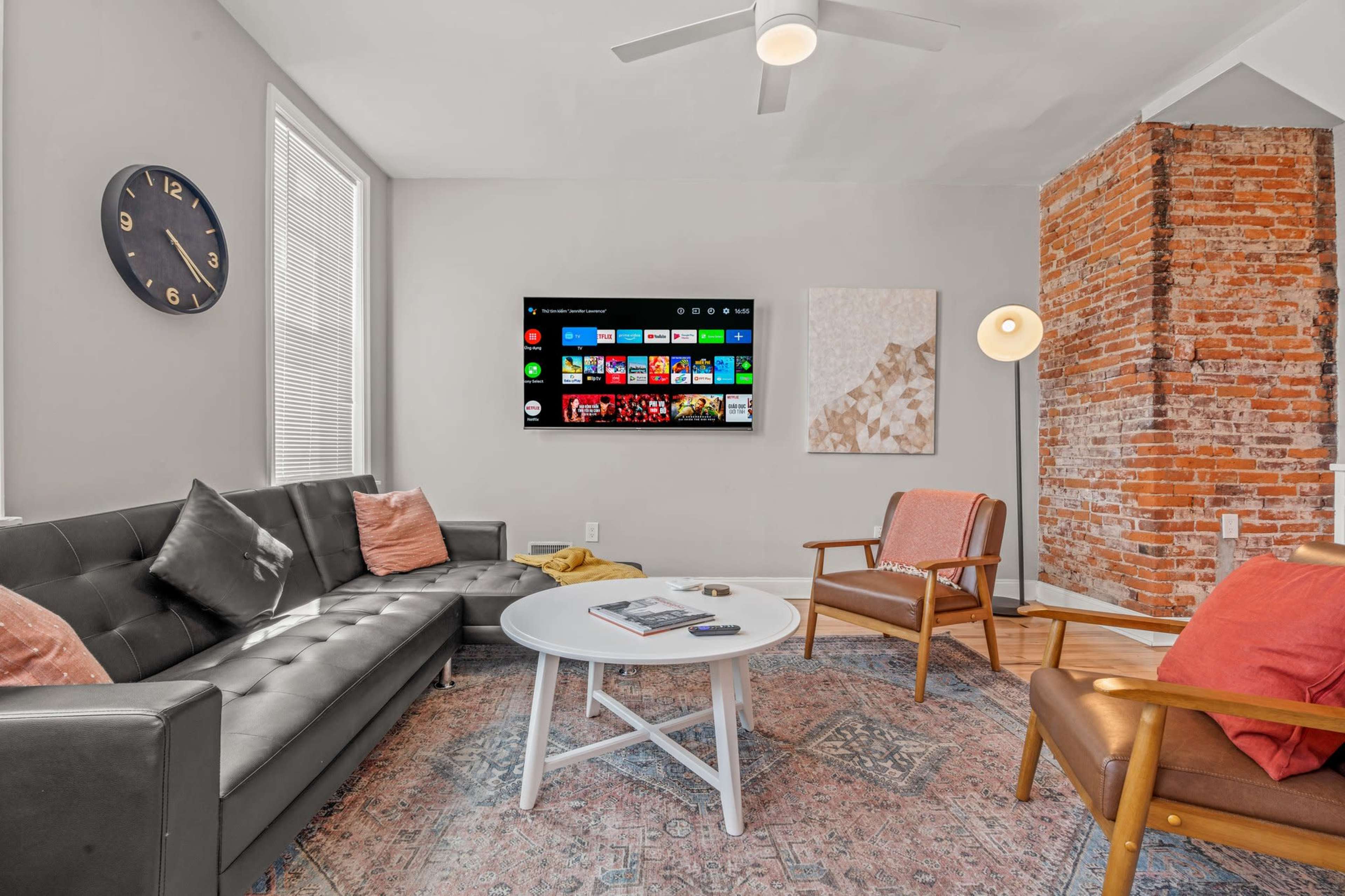 The living room features a modern television mounted on the wall above a light-colored coffee table, flanked by a dark couch and an armchair, with a brick accent wall in the background.
