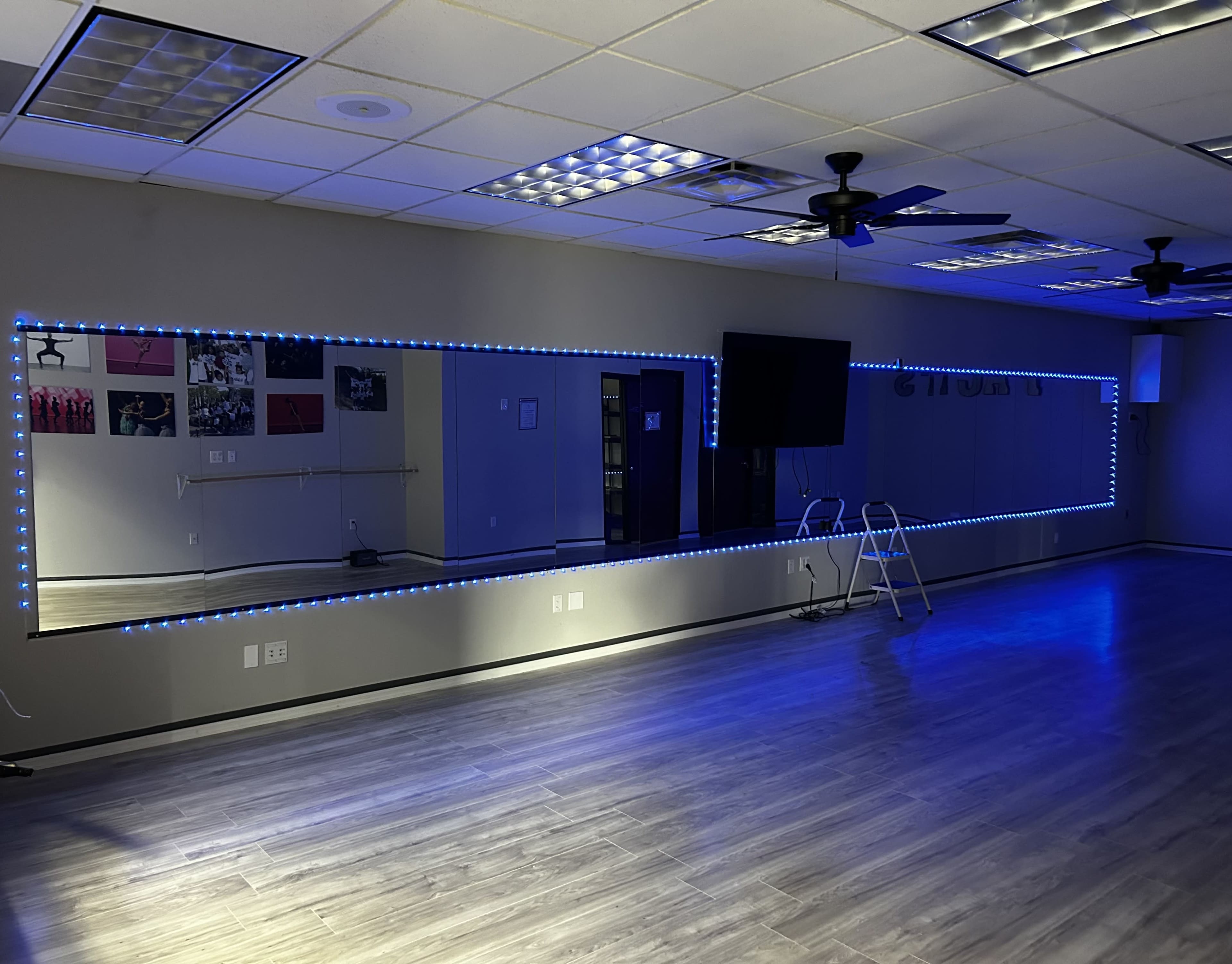 A spacious room features a long mirror framed with blue LED lights, wooden flooring, and a wall-mounted television.