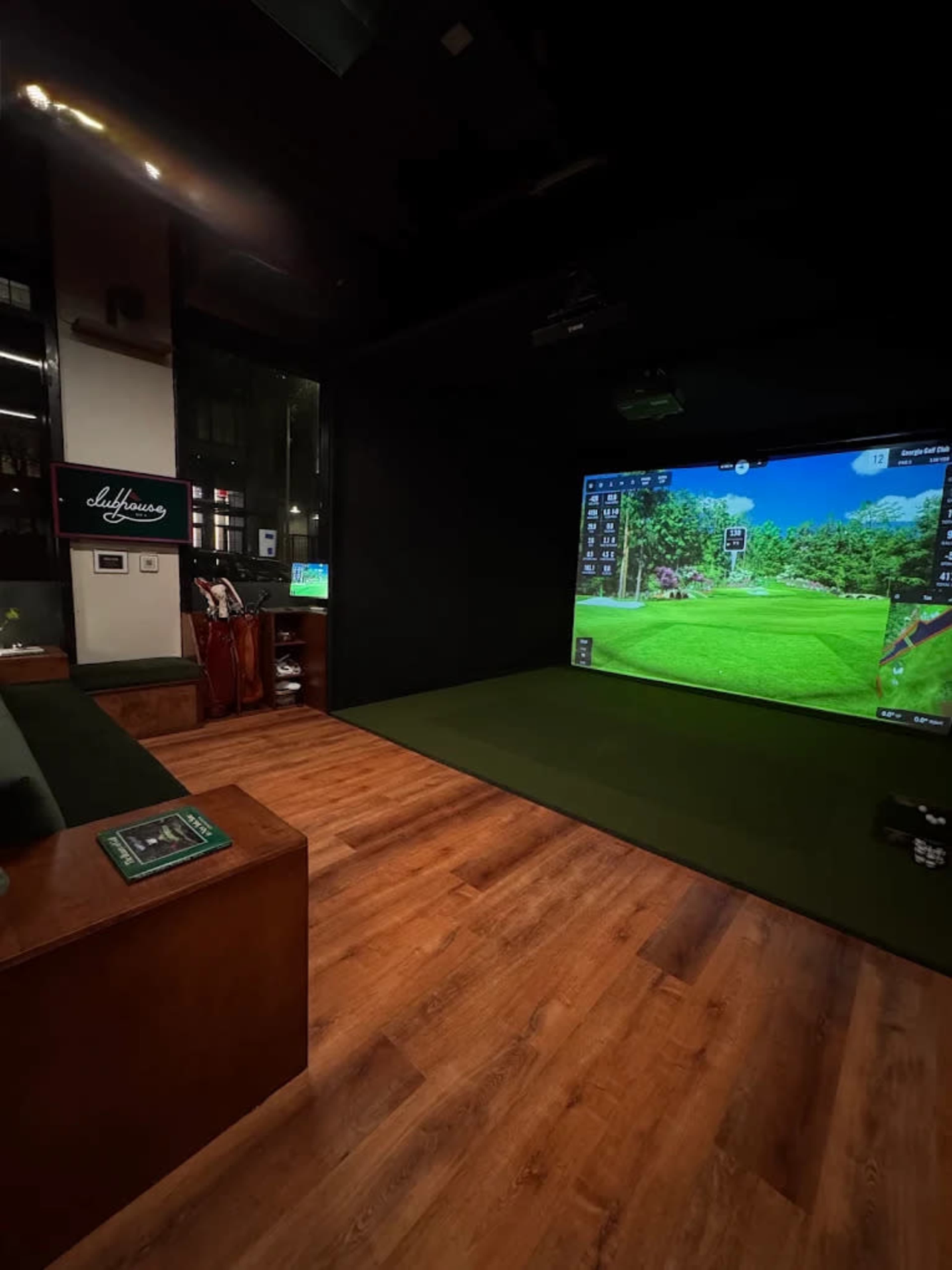 The image shows an indoor golf simulation room featuring a large screen displaying a golf course and a putting mat, with golf clubs and bags in the background.