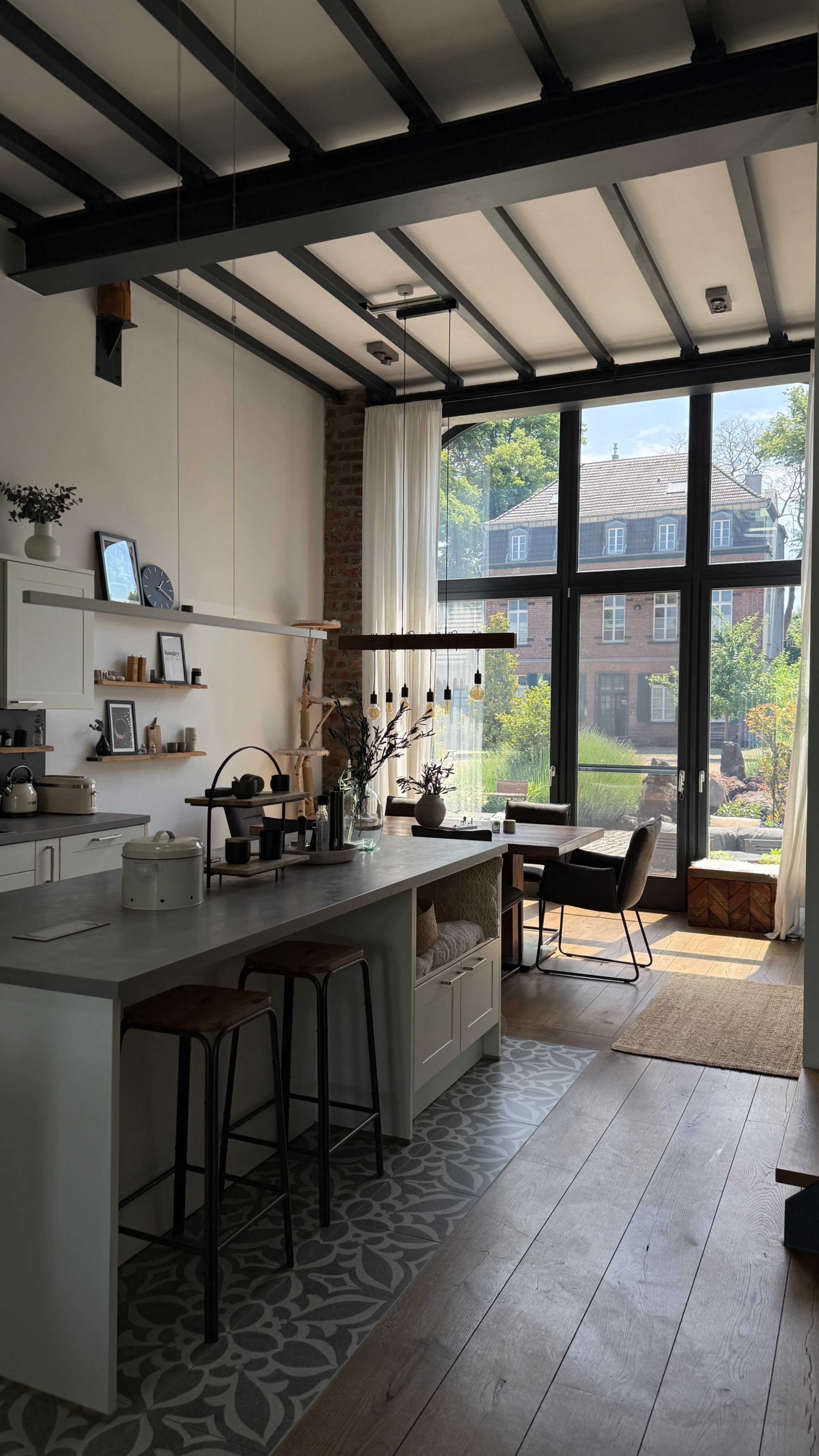 The image shows a modern kitchen with a central island, bar stools, and large windows that allow natural light to illuminate the space.