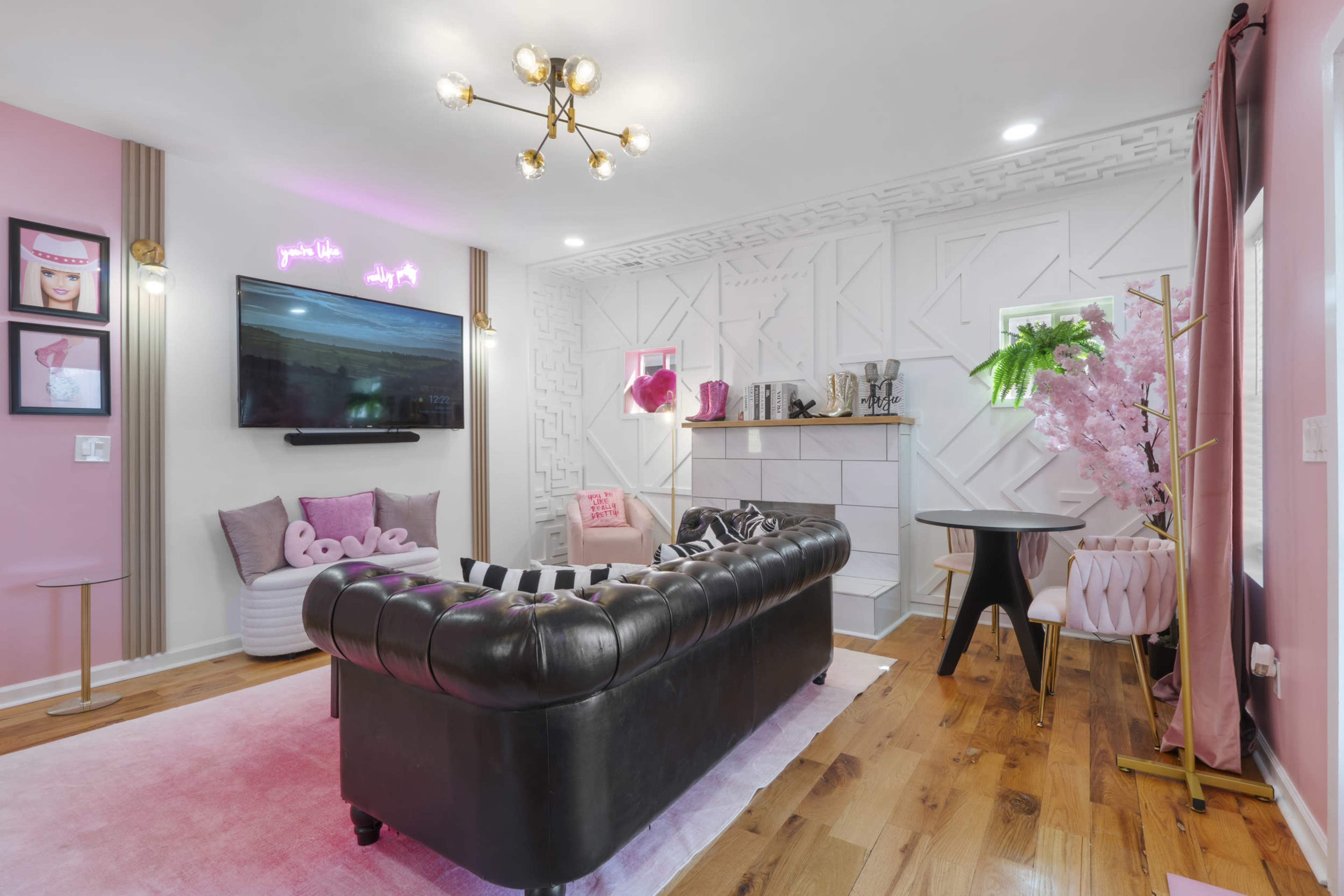 The image shows a modern living room with a black tufted sofa, pink decor elements, and a wall-mounted TV above a stylish white fireplace.