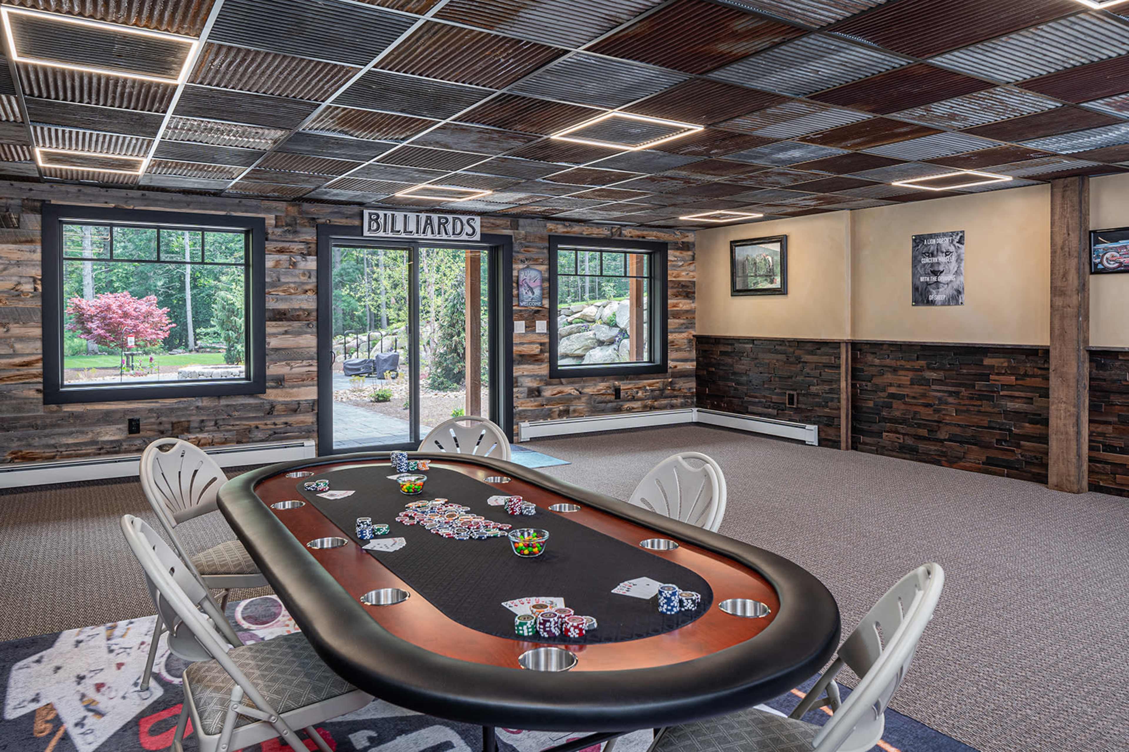 A game room featuring a poker table surrounded by chairs, with large windows showing a view of greenery outside.