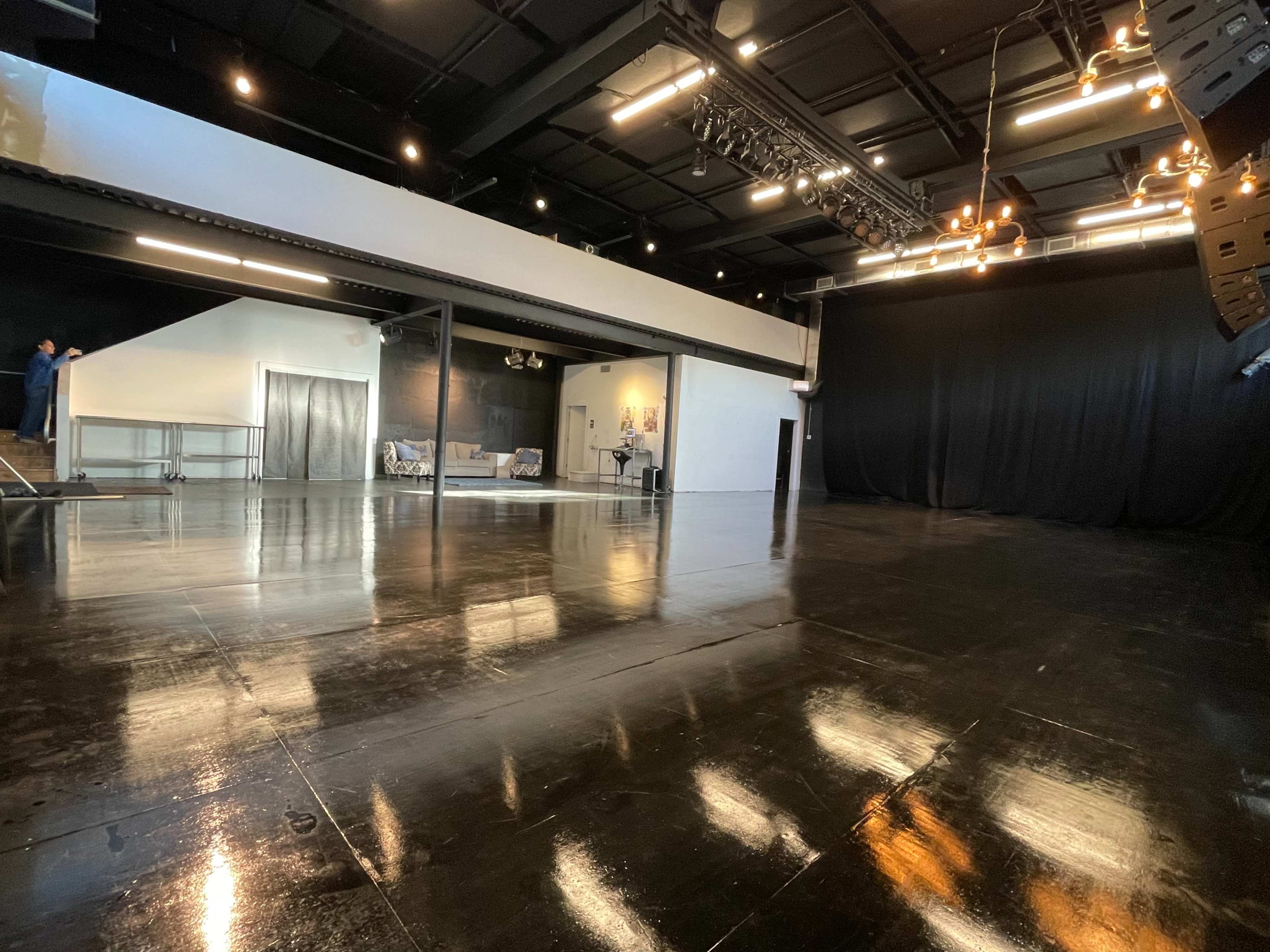 A spacious, polished performance studio features a large, empty floor, black curtains, and a few people in the background.