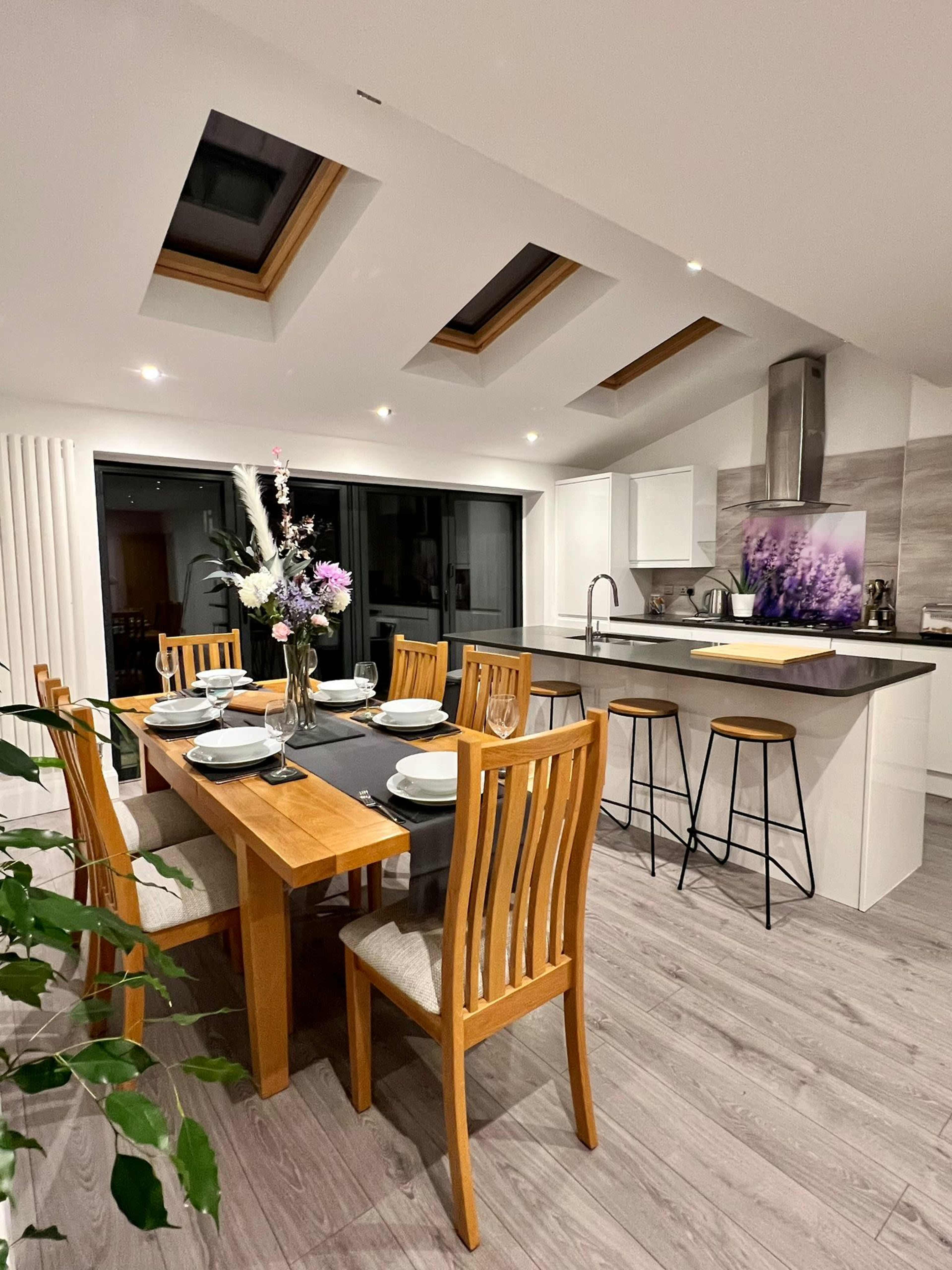 A modern kitchen features a dining table with eight chairs, skylights above, a counter with bar stools, and a floral centerpiece.