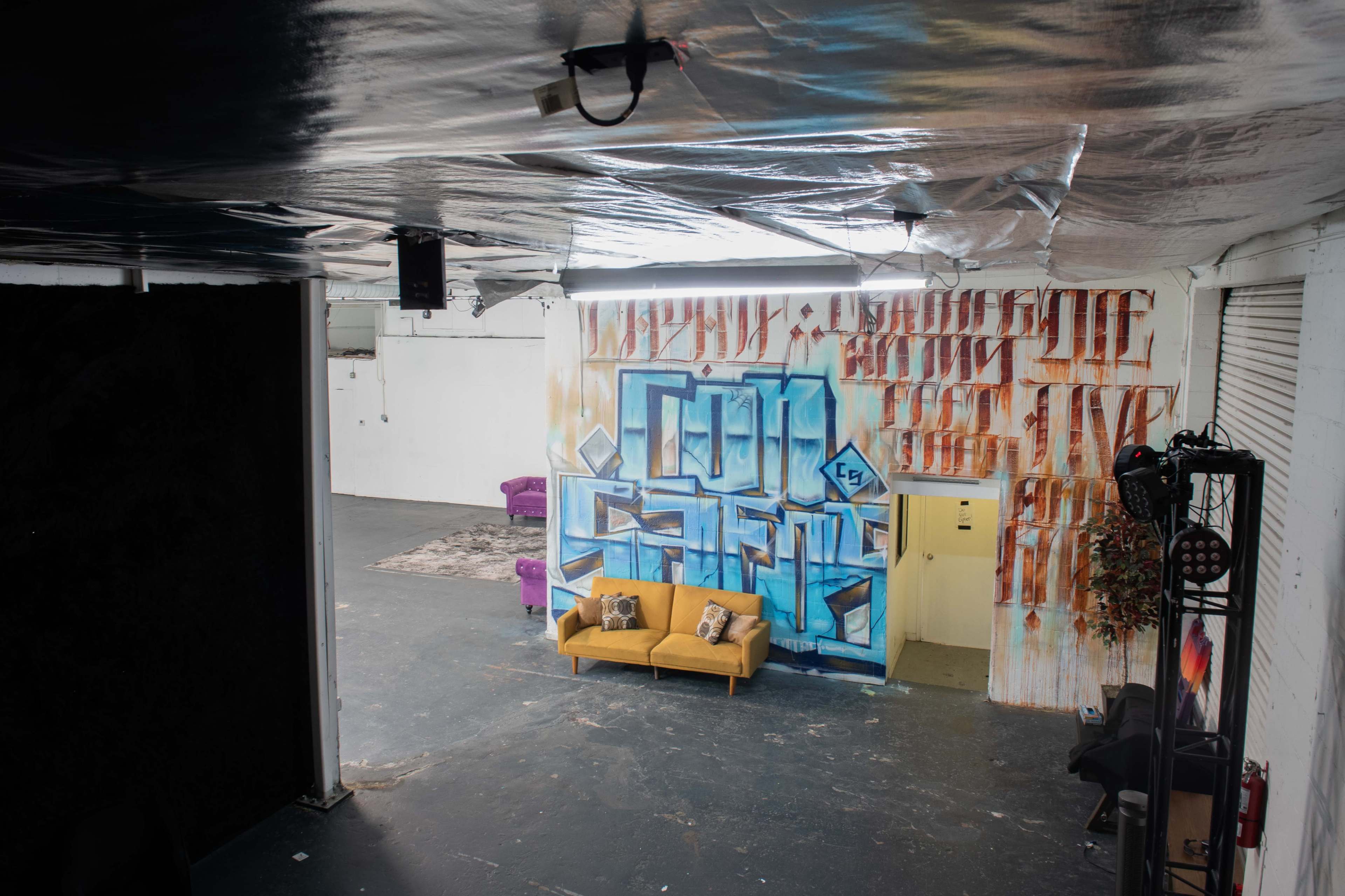 An industrial-style interior with a yellow sofa in front of a large blue graffiti mural, complemented by a purple armchair and various textures on the walls.