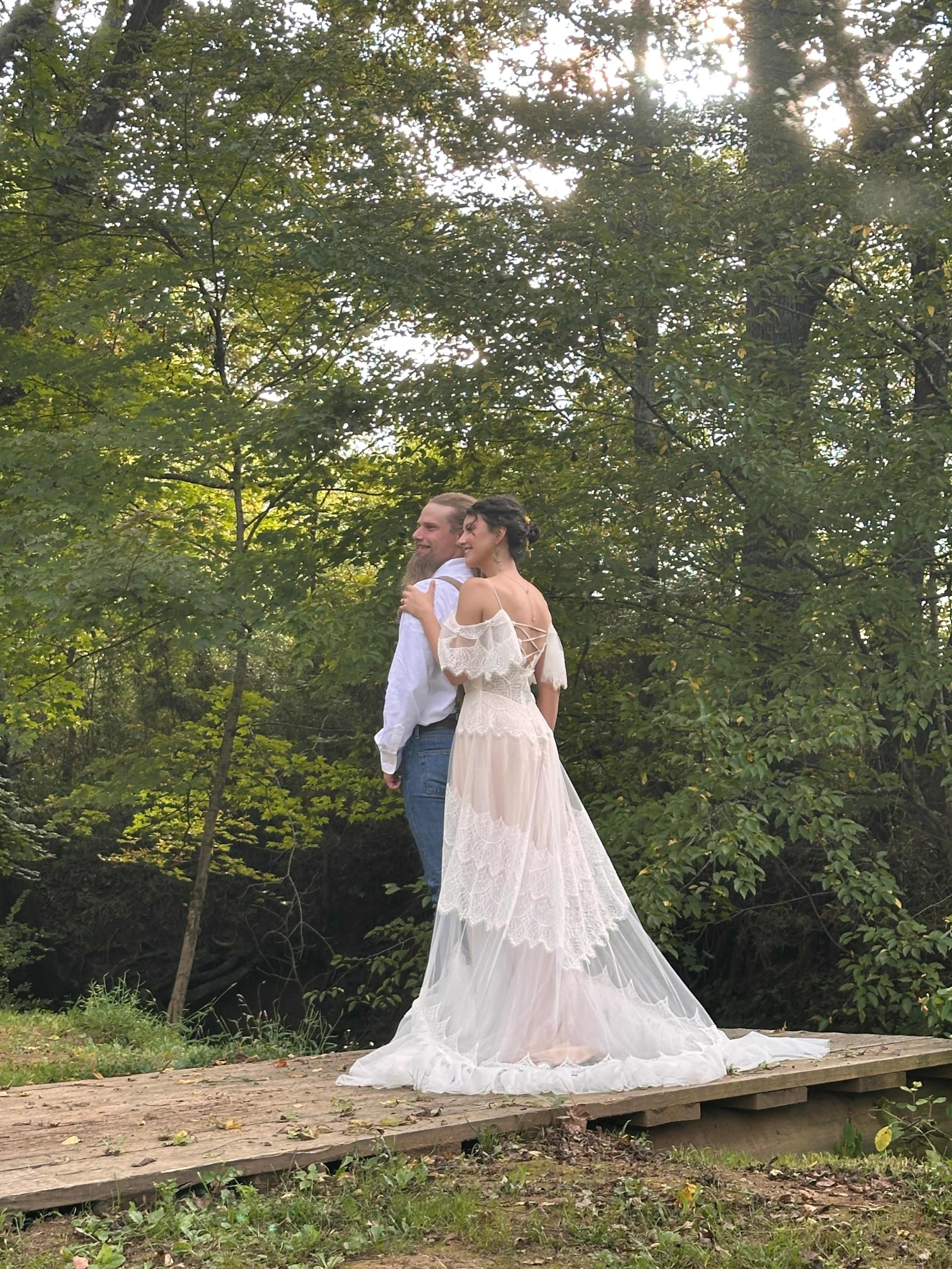 A couple stands together on a wooden path in a forested area, with the woman in a flowing wedding dress and the man in a white shirt and jeans.
