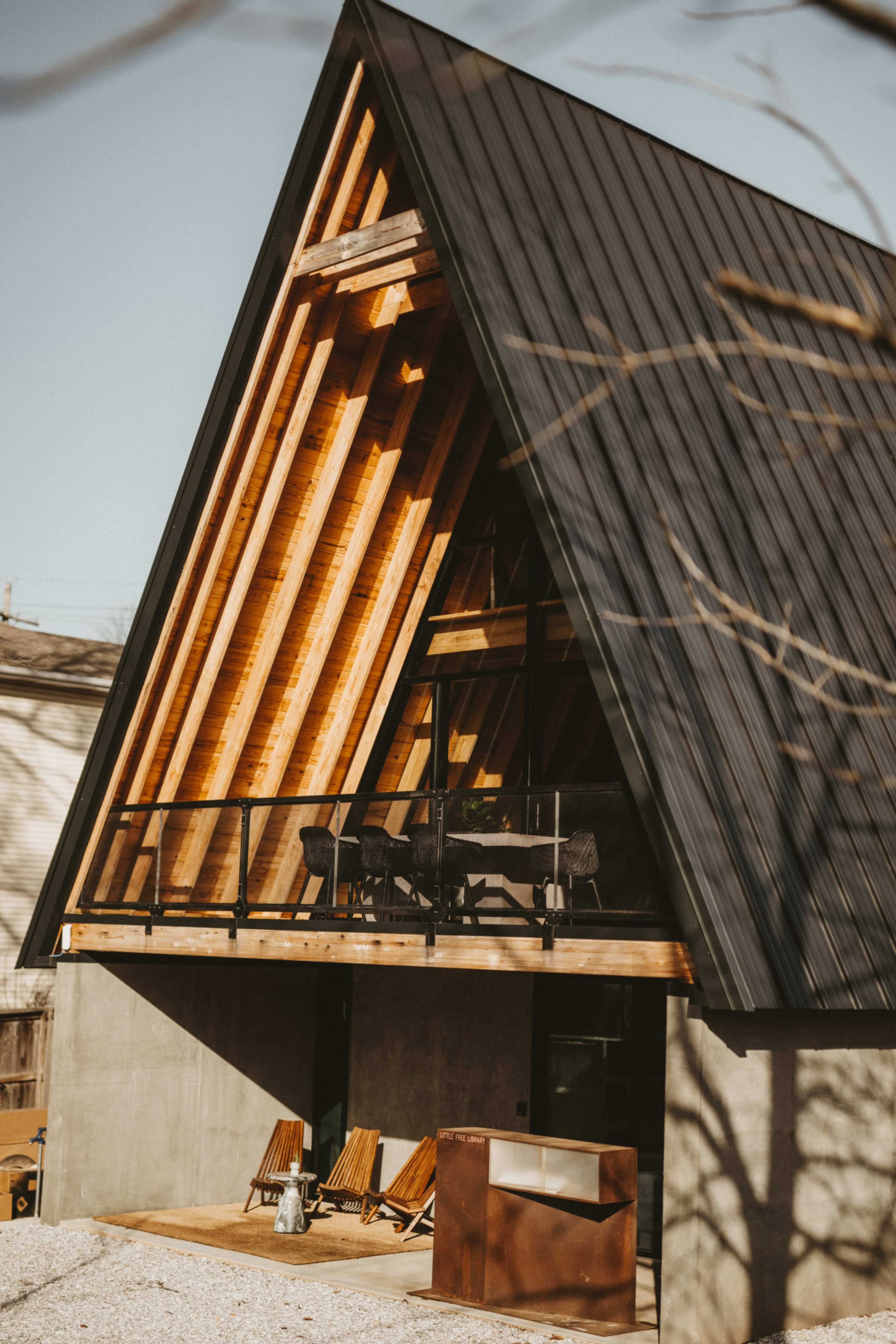 A modern A-frame house with a triangular roof, wooden beams, and a concrete base, featuring a balcony and patio furniture in the foreground.