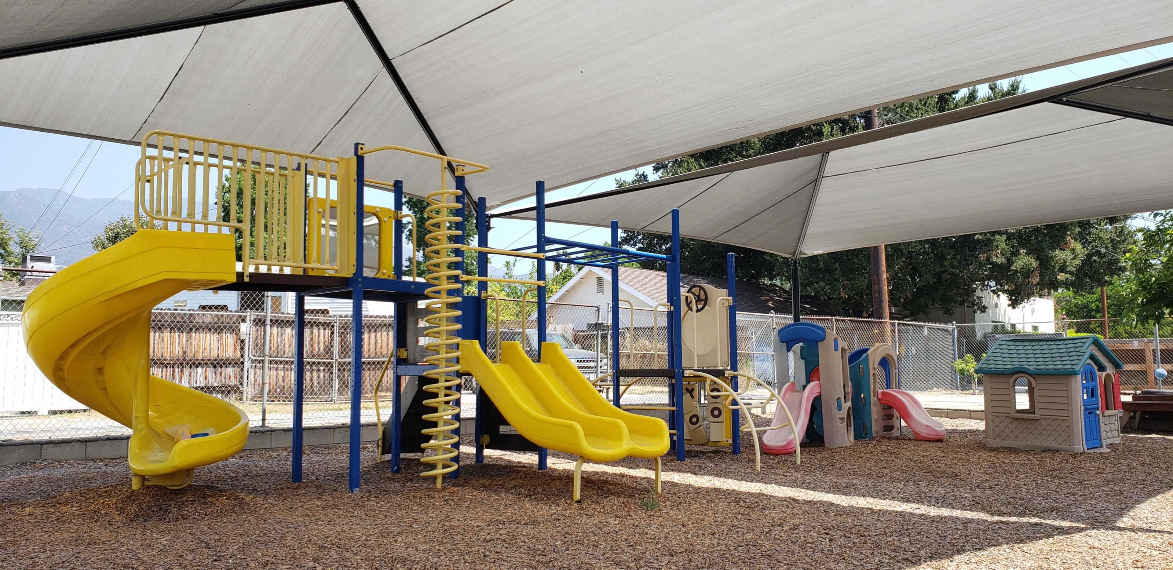 The image shows a covered playground featuring various slides, climbing structures, and a small playhouse set on a bed of wood chips.