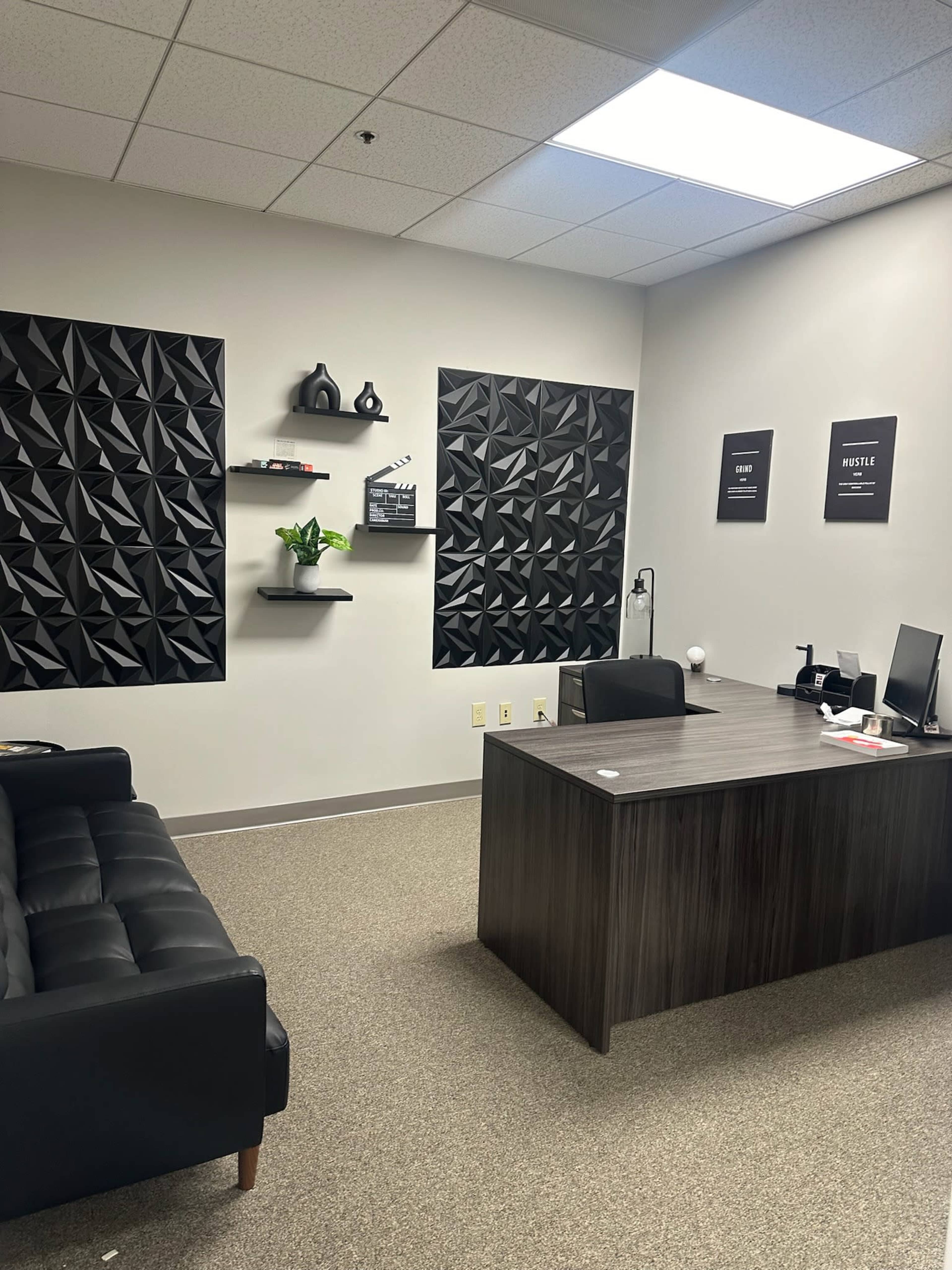 The image shows a modern office space featuring a dark couch, a desk with a computer, and textured black wall decor.