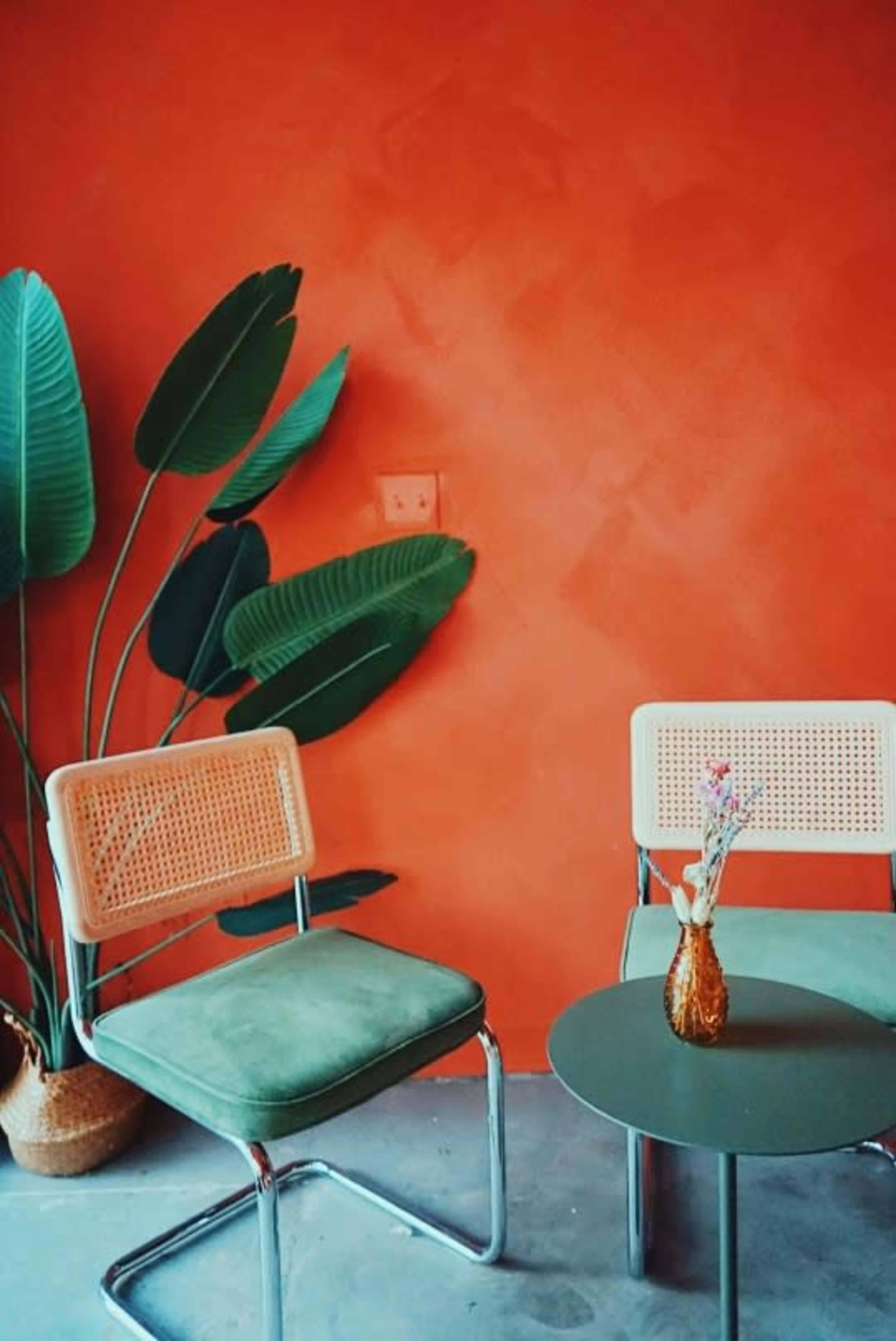 Two chairs beside a green table with a vase containing flowers against an orange wall decorated with large green leaves.
