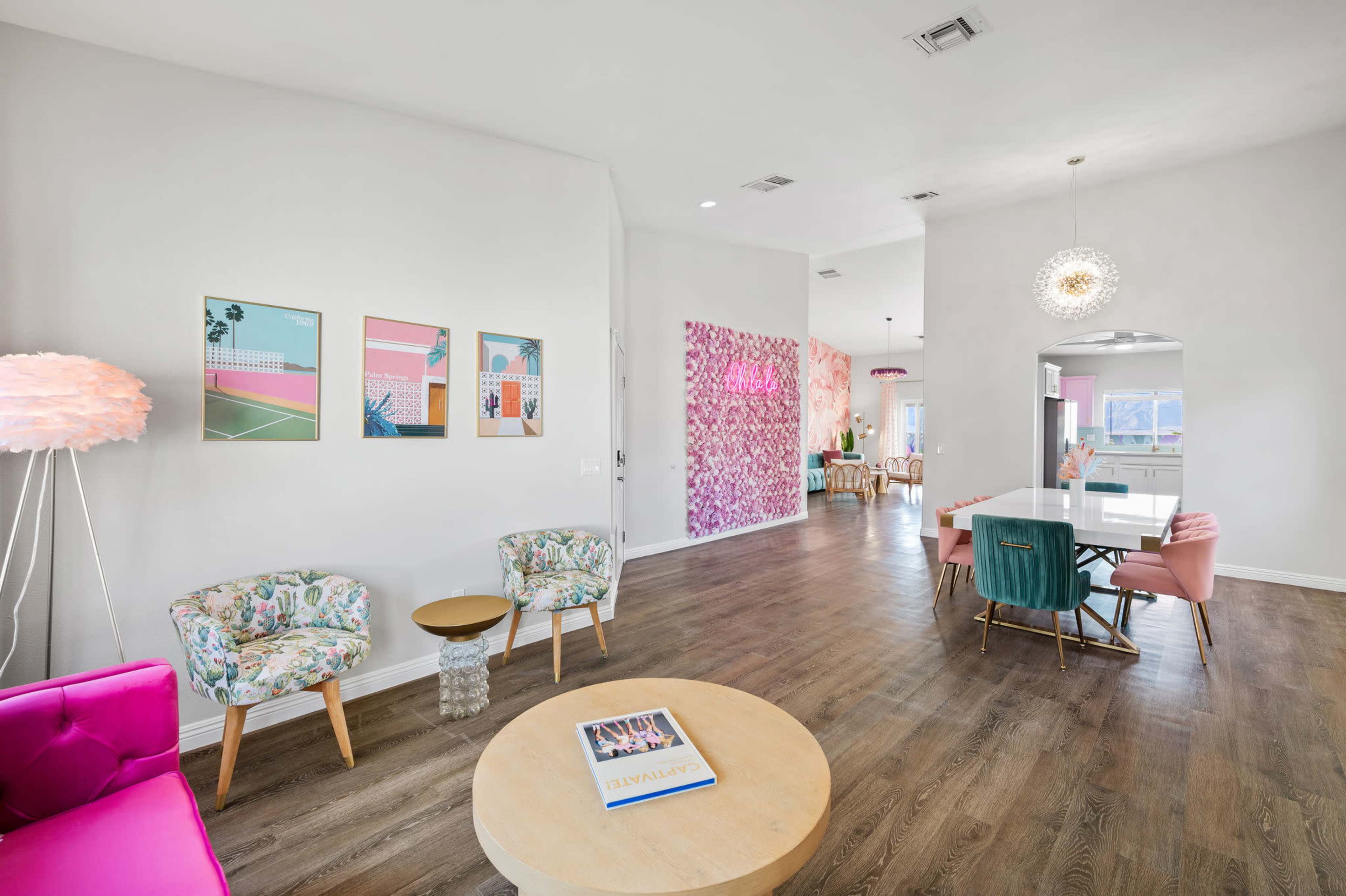 The room features a living area with floral-patterned chairs, a pink sofa, and a round wooden coffee table, leading into a dining space with colorful decor and a light fixture.