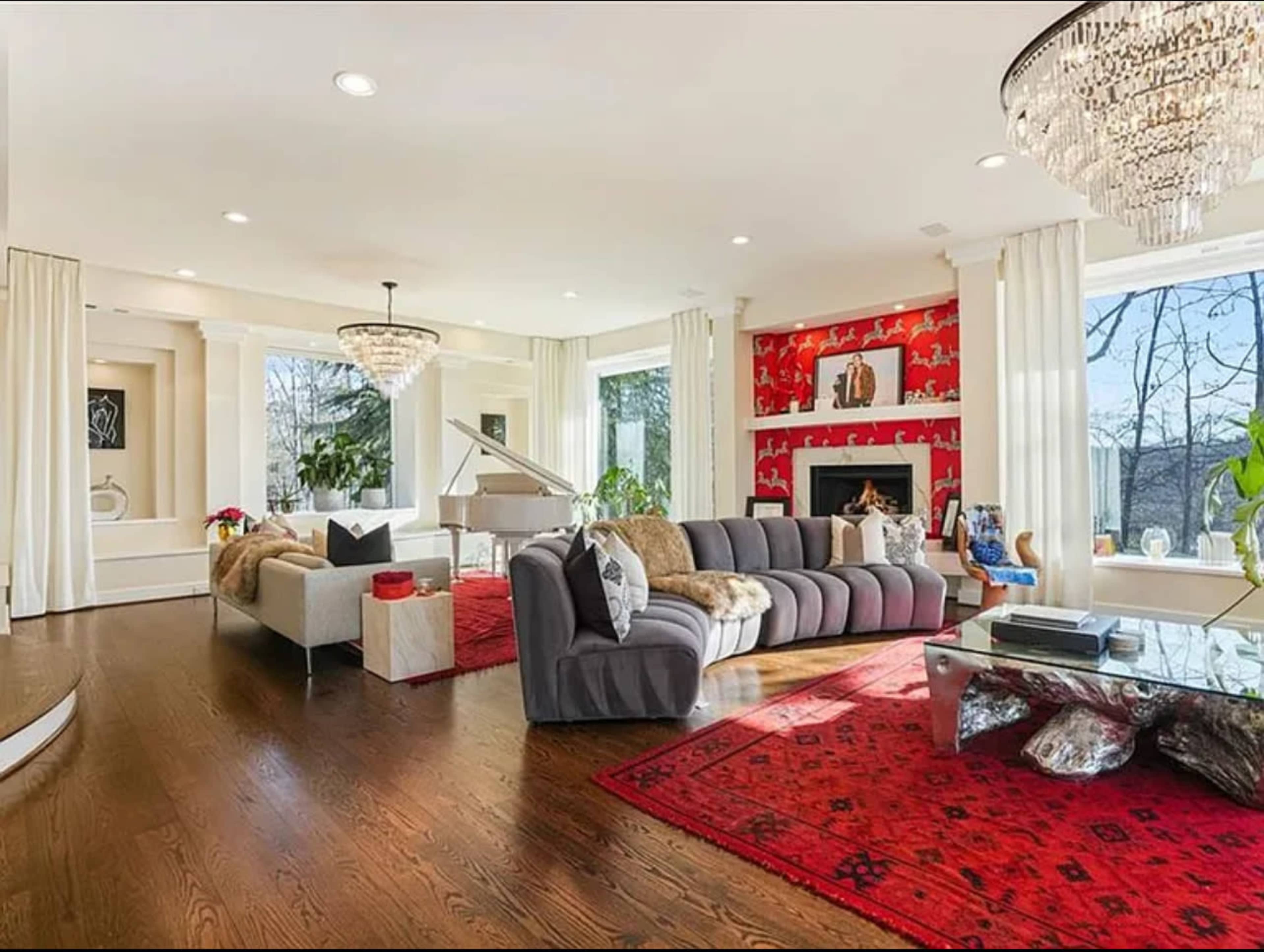 The image depicts a modern living room featuring a large gray sectional sofa, a glass coffee table, a piano, and a vibrant red accent wall, with large windows letting in natural light.