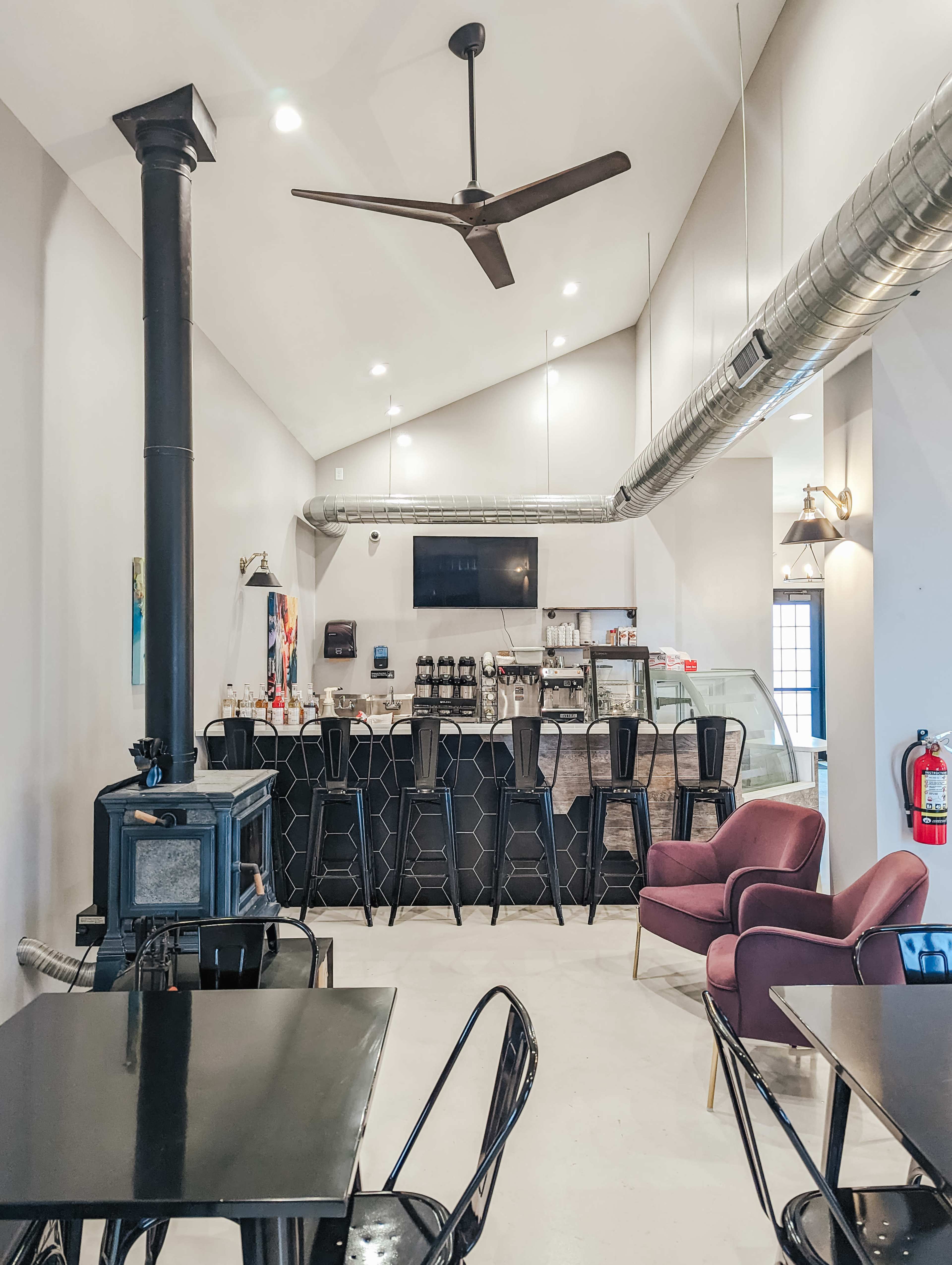 The interior of a café features a counter with seating, a stove in the corner, and a high ceiling with ceiling fans and industrial-style lighting.