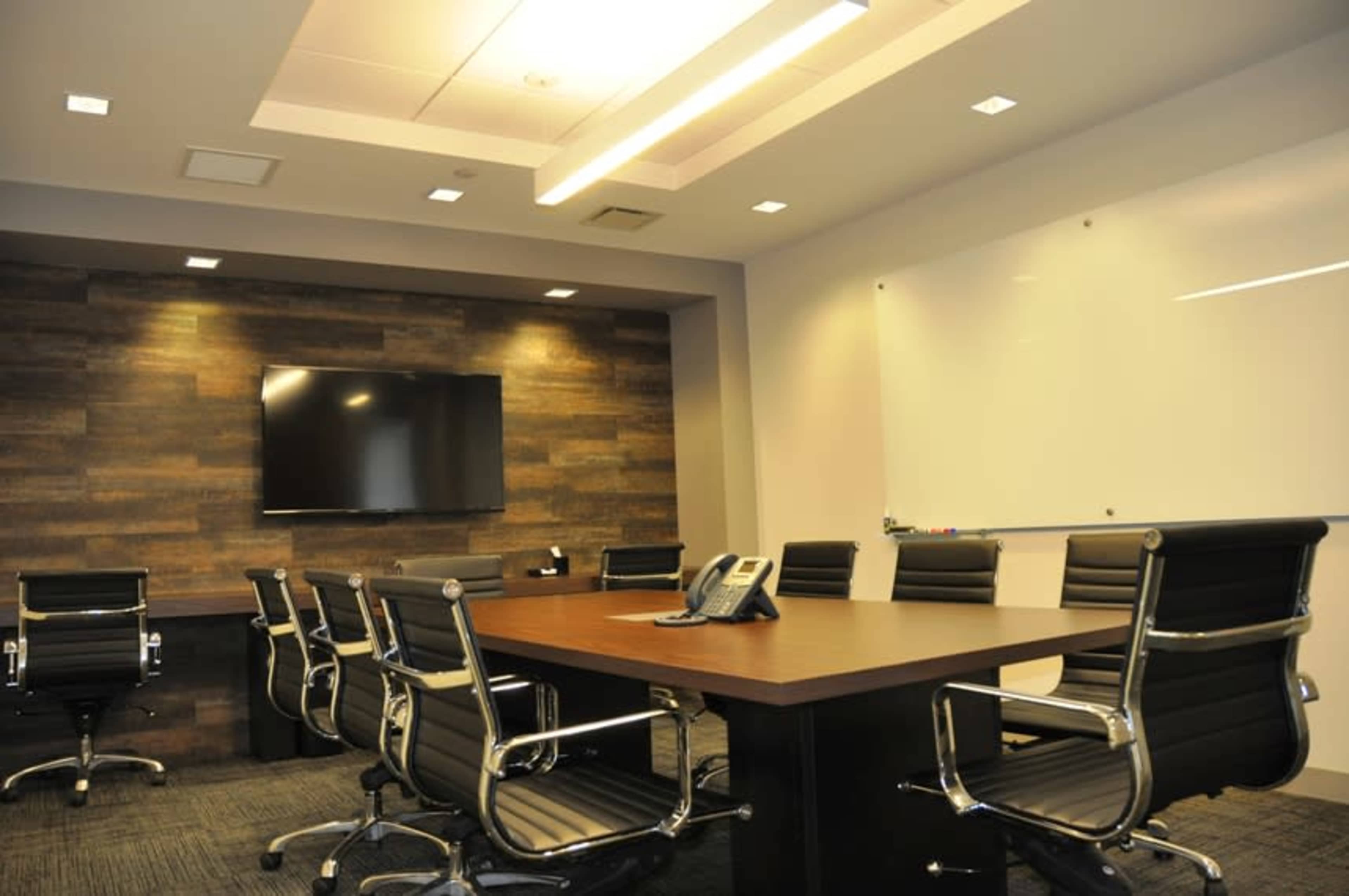 The image shows a modern conference room with a large table, ergonomic chairs, a wall-mounted TV, and a whiteboard.