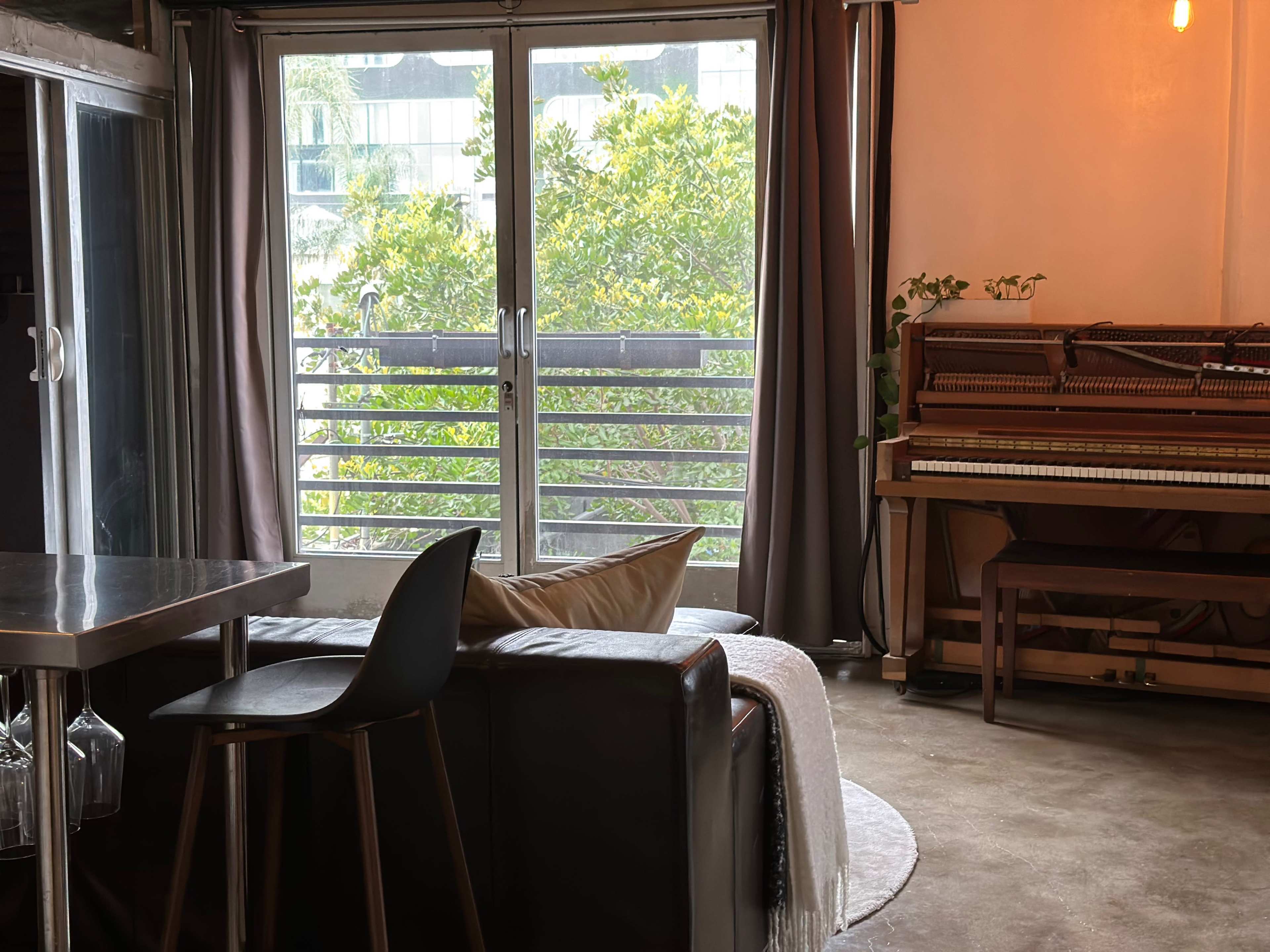 A modern living room with a black sofa, a dining area with a metal table, and a piano next to a large window that offers a view of greenery outside.