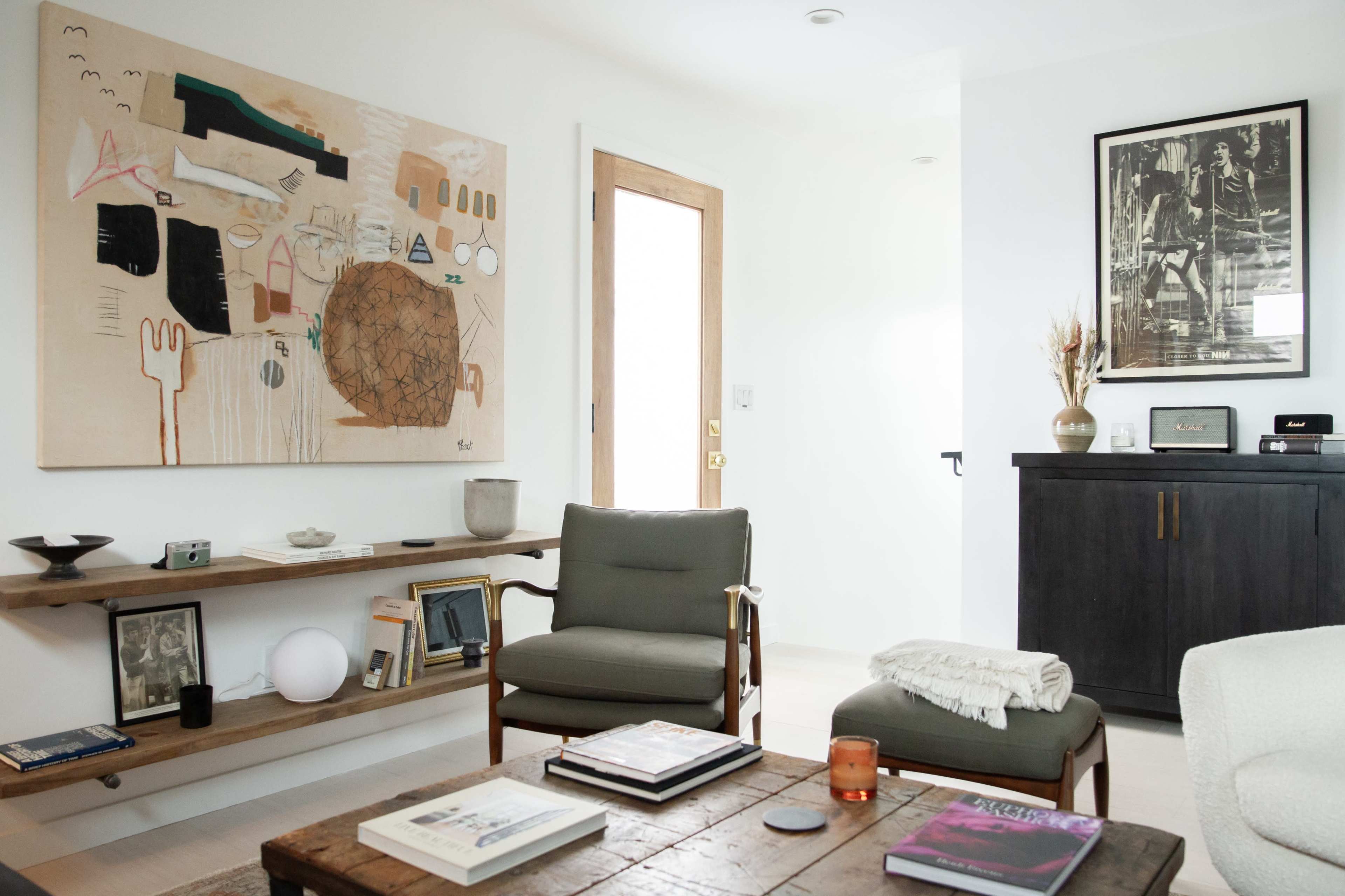 A modern living room features a textured painting on the wall, a wooden coffee table with books, two chairs, and a dark cabinet.