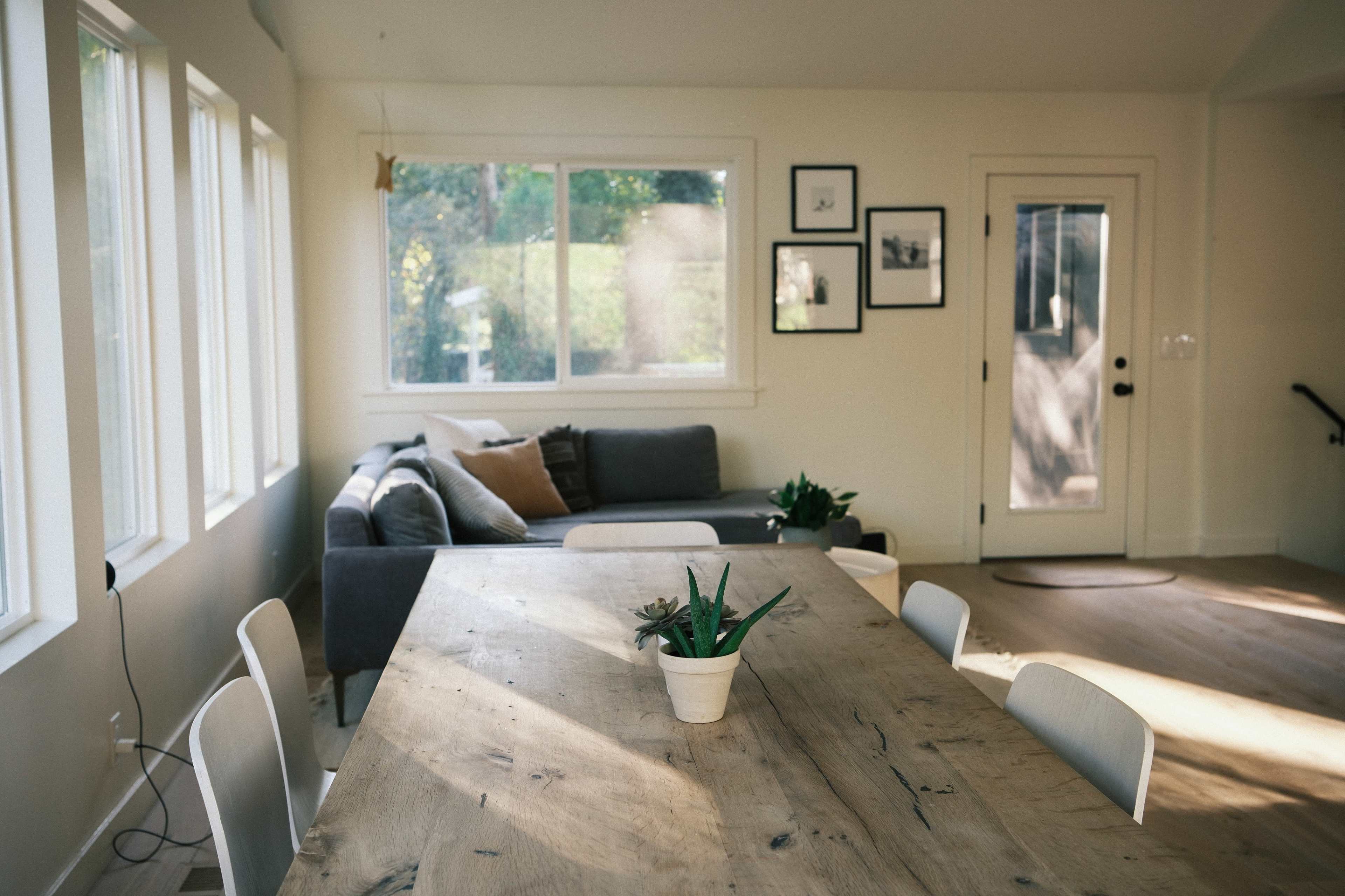The image shows a light-filled living space featuring a wooden dining table with a potted plant, a gray sofa, and several framed pictures on the walls.